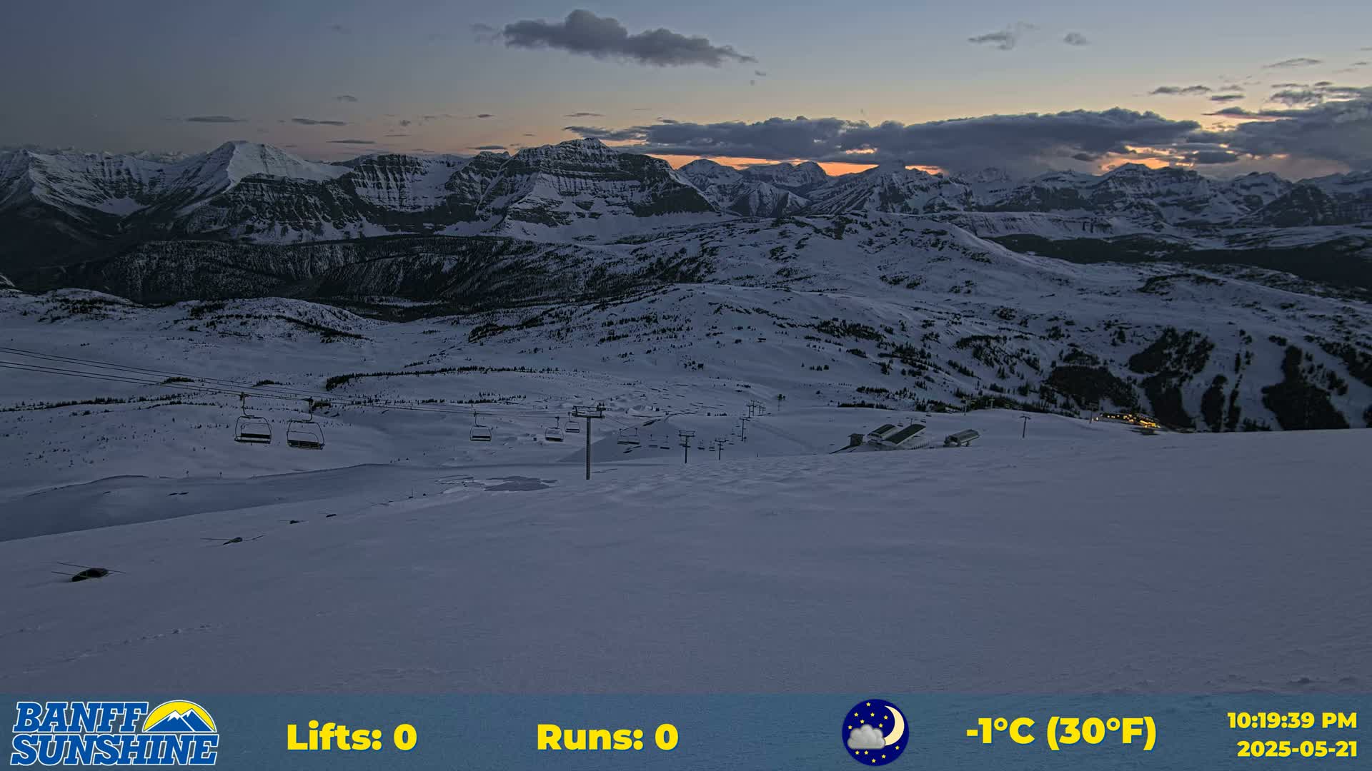 A snow-covered ski resort at dusk, with several chairlifts visible against a backdrop of mountains under a partly cloudy sky.