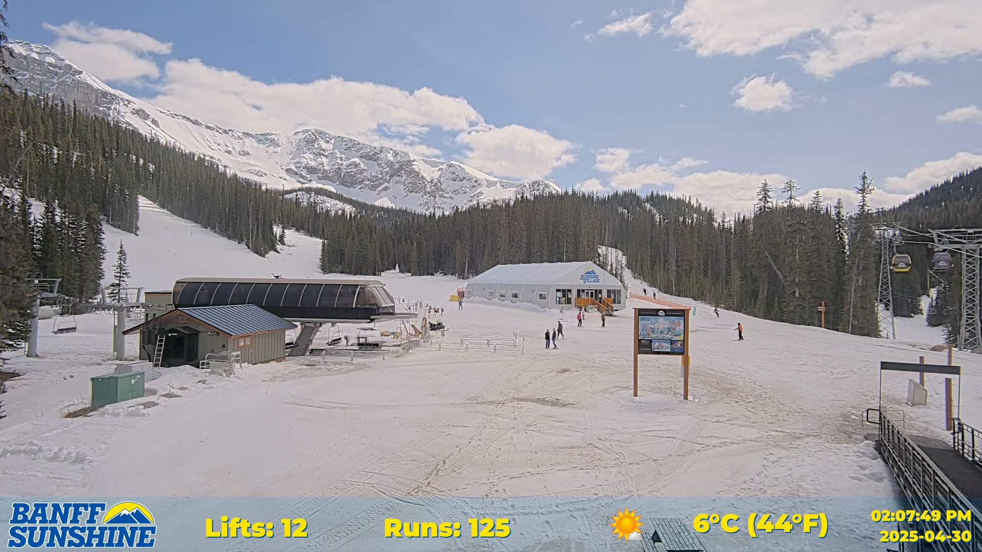 A sunny ski resort with snow-covered slopes, a chairlift, a small building, and a few people scattered about, all set against a backdrop of snow-capped mountains.