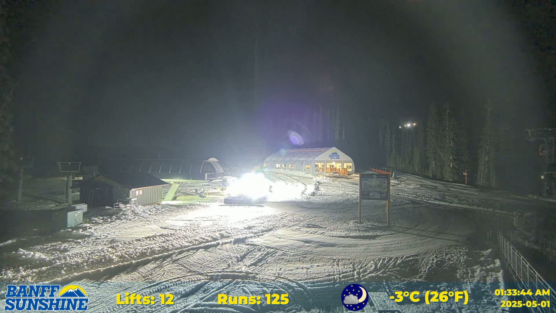 A snow-covered ski resort at night, illuminated by bright lights, with a building and ski lifts visible under a partly cloudy sky.
