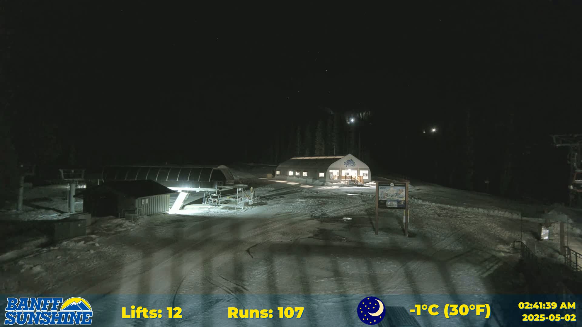 A nighttime view of a snow-covered ski resort with several buildings illuminated, under a clear, starry sky and a temperature of -1°C (30°F).