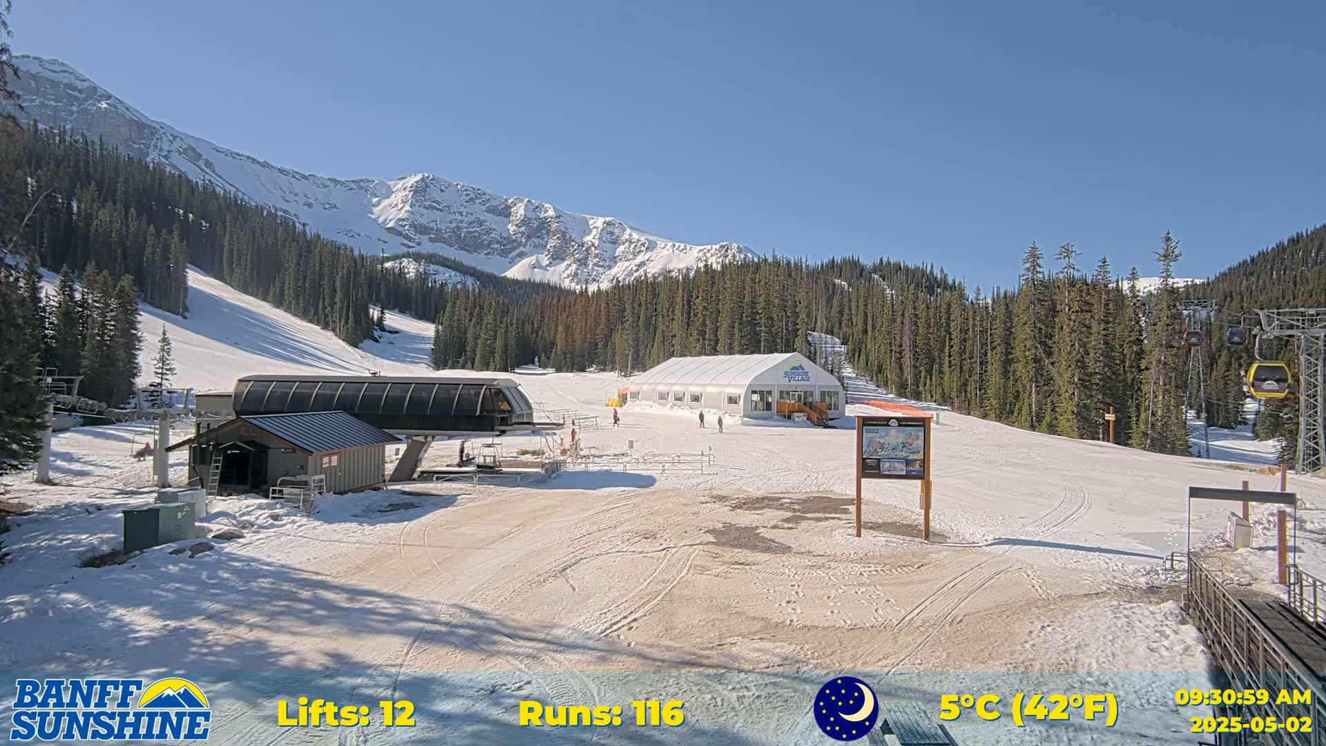 A sunny ski resort scene shows snow-covered slopes, ski lifts, a small building, and a large tent, all nestled among evergreen trees and snow-capped mountains in the background.