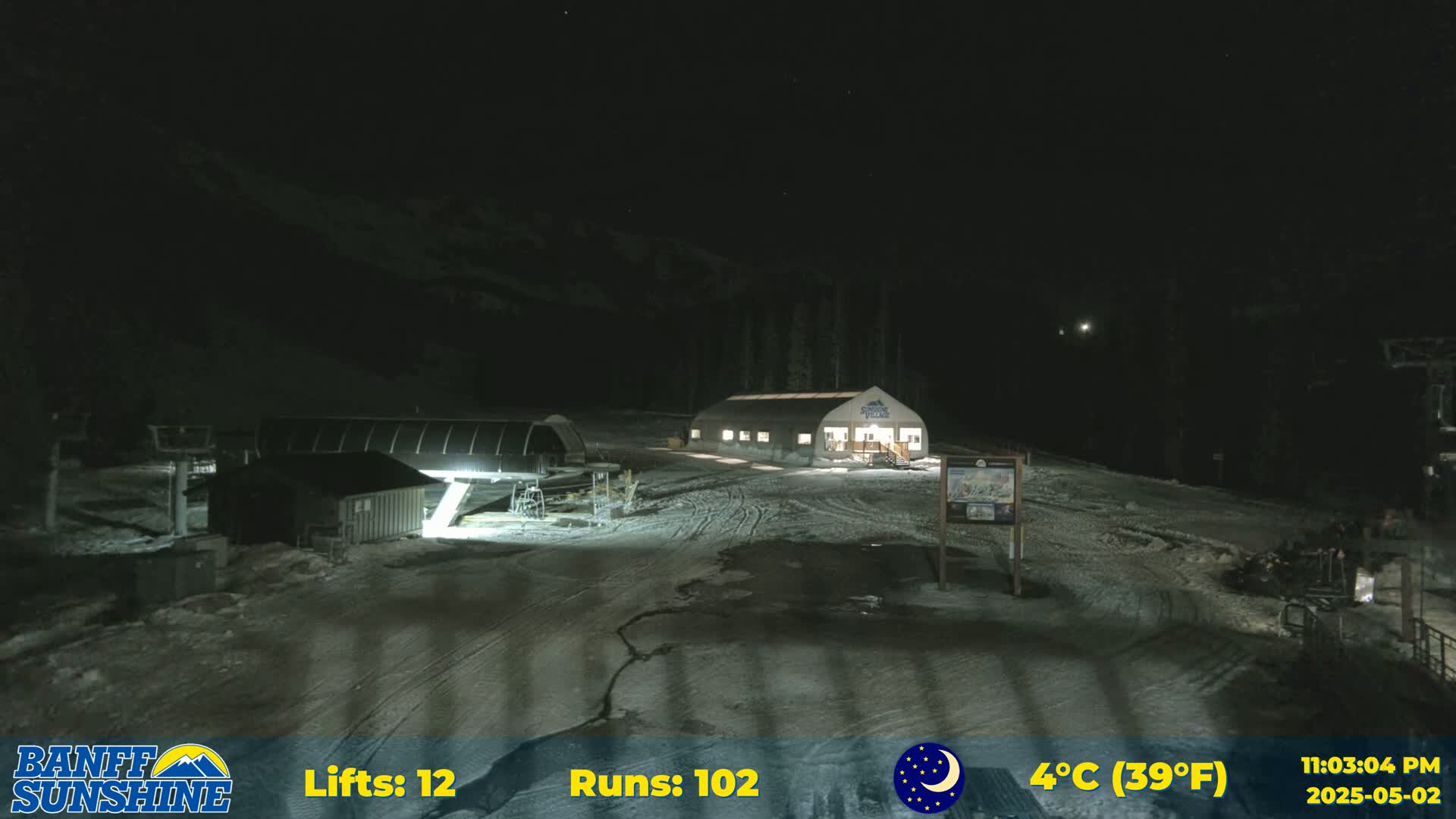 A nighttime view of a snow-covered ski resort with a building and ski lifts, under a clear, dark sky with a temperature of 4°C (39°F).