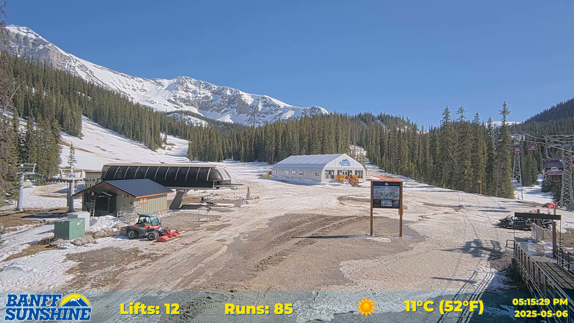 A sunny ski resort base area with patchy snow, a chairlift, and a small building, situated against a backdrop of snow-capped mountains and evergreen trees.
