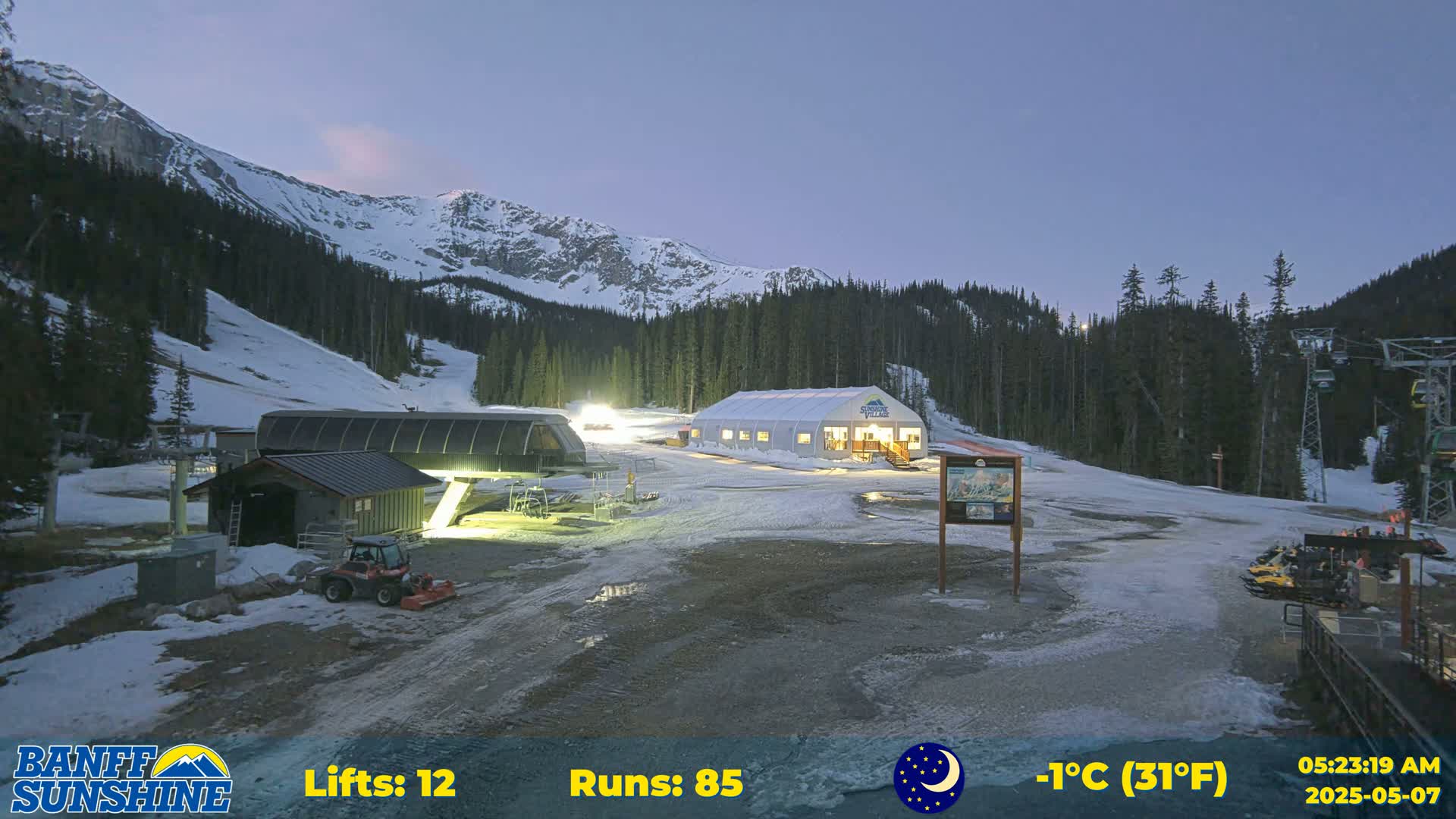 A ski resort at dusk with snow-covered ground, illuminated buildings, and a mountain range in the background under a twilight sky.