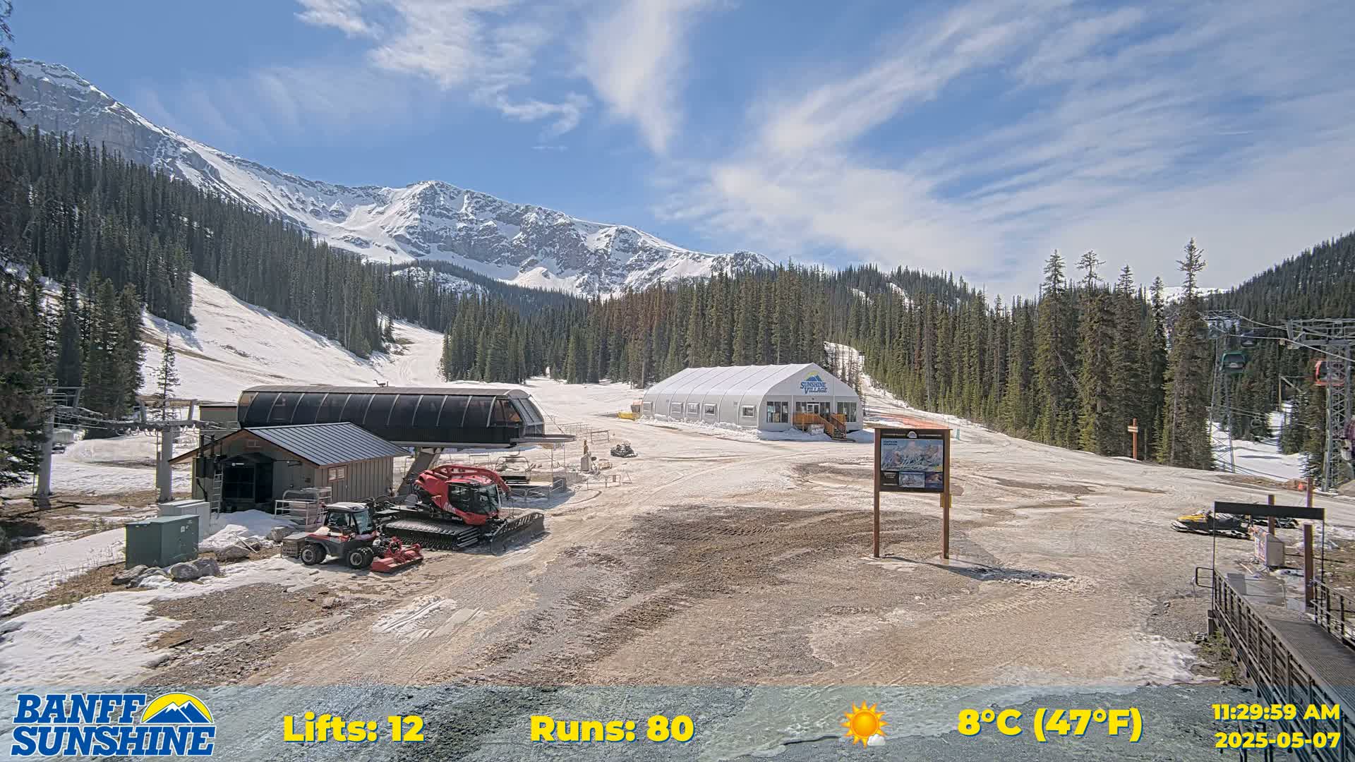 A ski resort base area with patchy snow, a large white tent, ski lifts, and snow grooming machinery, is situated in front of a mountain range under a mostly sunny sky.