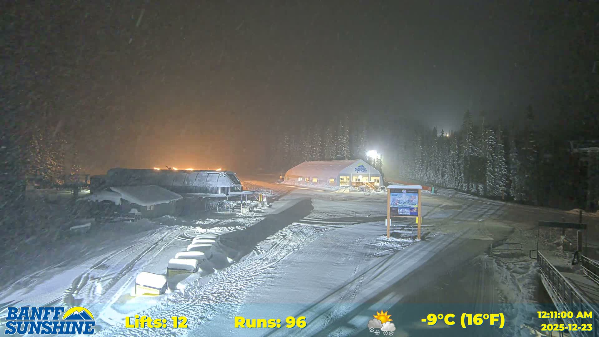 A ski resort base area with patchy snow, a large white tent, ski lifts, and snow grooming machinery, is situated in front of a mountain range under a mostly sunny sky.