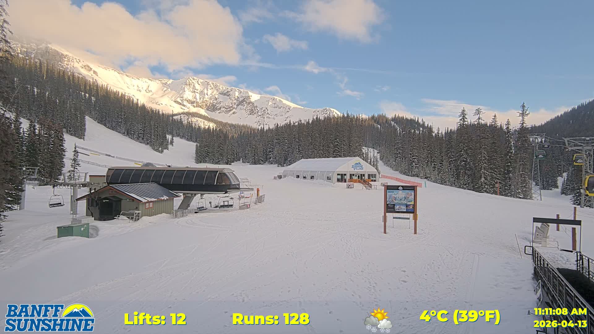 A ski resort base area with patchy snow, a large white tent, ski lifts, and snow grooming machinery, is situated in front of a mountain range under a mostly sunny sky.