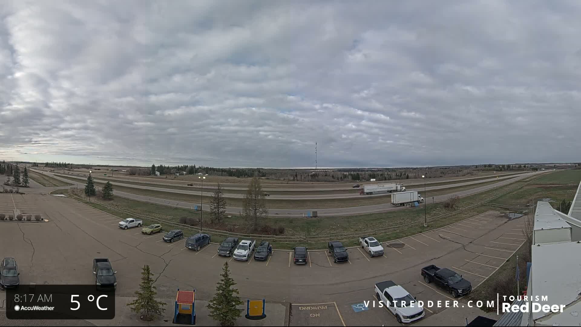 A mostly cloudy sky hangs over a highway with several vehicles, including semi-trucks, and a parking lot with numerous cars, all under a 5°C temperature.
