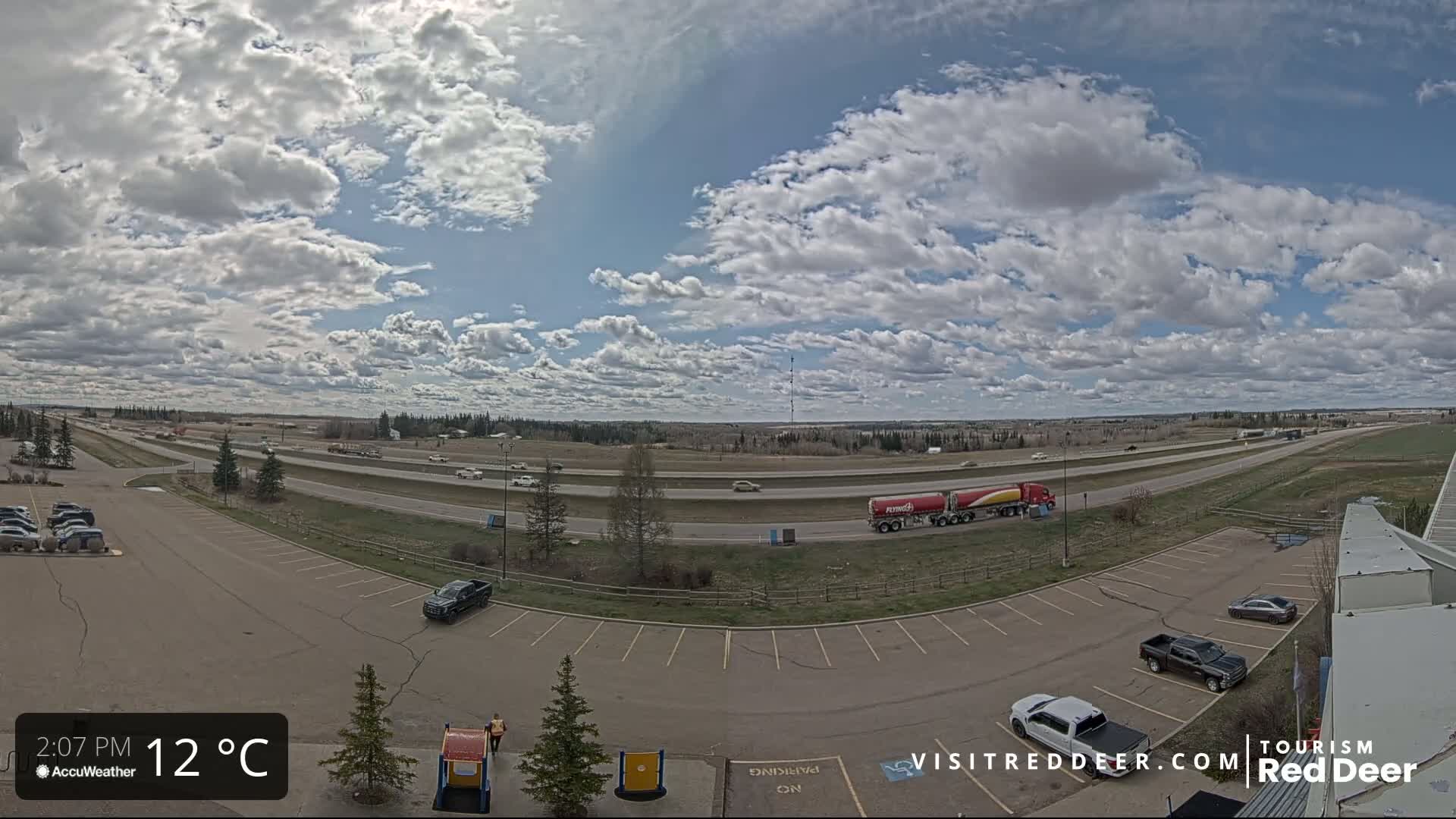 A partly cloudy sky overlooks a highway with moderate traffic and an empty parking lot containing several vehicles.