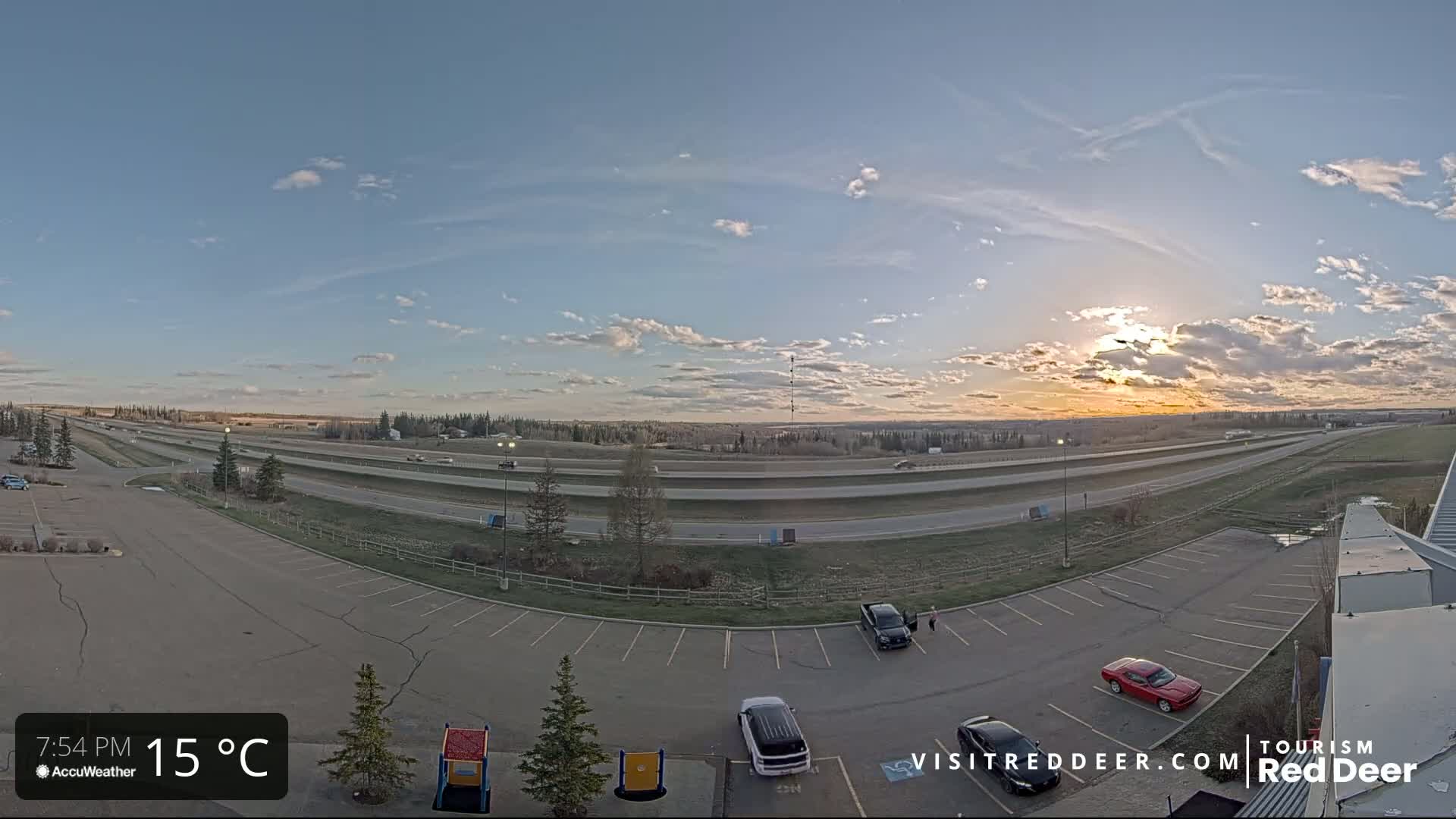 A mostly sunny, partly cloudy evening scene shows a highway with sparse traffic and an empty parking lot with a few cars, all viewed from an elevated position.