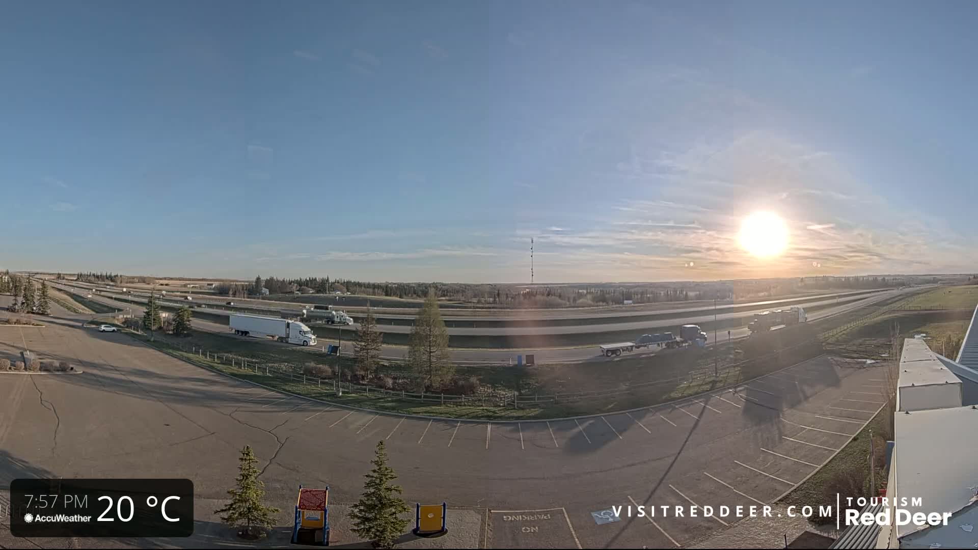 A mostly sunny, 20°C afternoon scene shows a highway with several vehicles, including semi-trucks, and an empty parking lot with a small playground next to it.