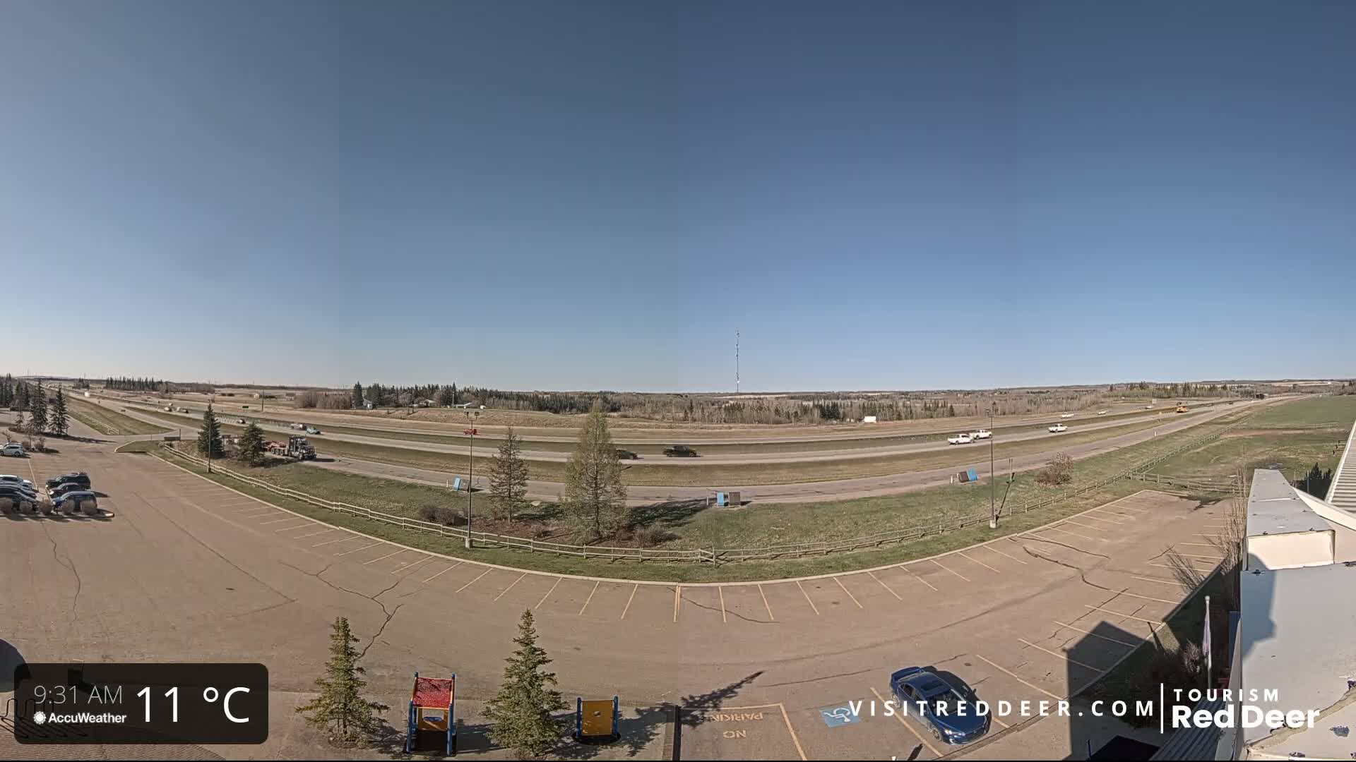 A mostly empty parking lot overlooks a multi-lane highway with sparse traffic under a clear blue sky on a sunny 11°C day.