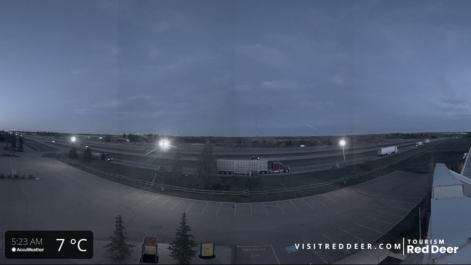 An empty parking lot at 7°C under a twilight sky overlooks a highway with several semi-trucks.