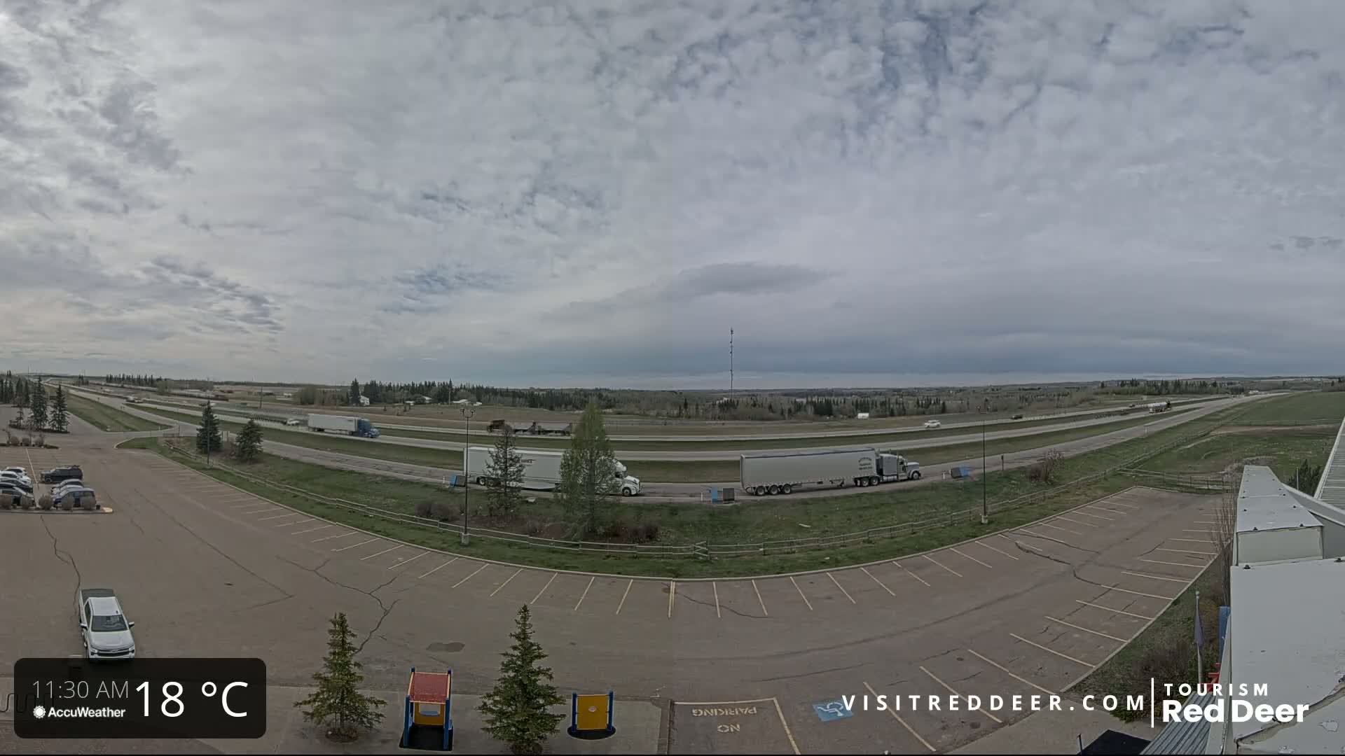 A mostly cloudy day shows a highway with several semi-trucks driving on it, a large parking lot, and some greenery in the distance.