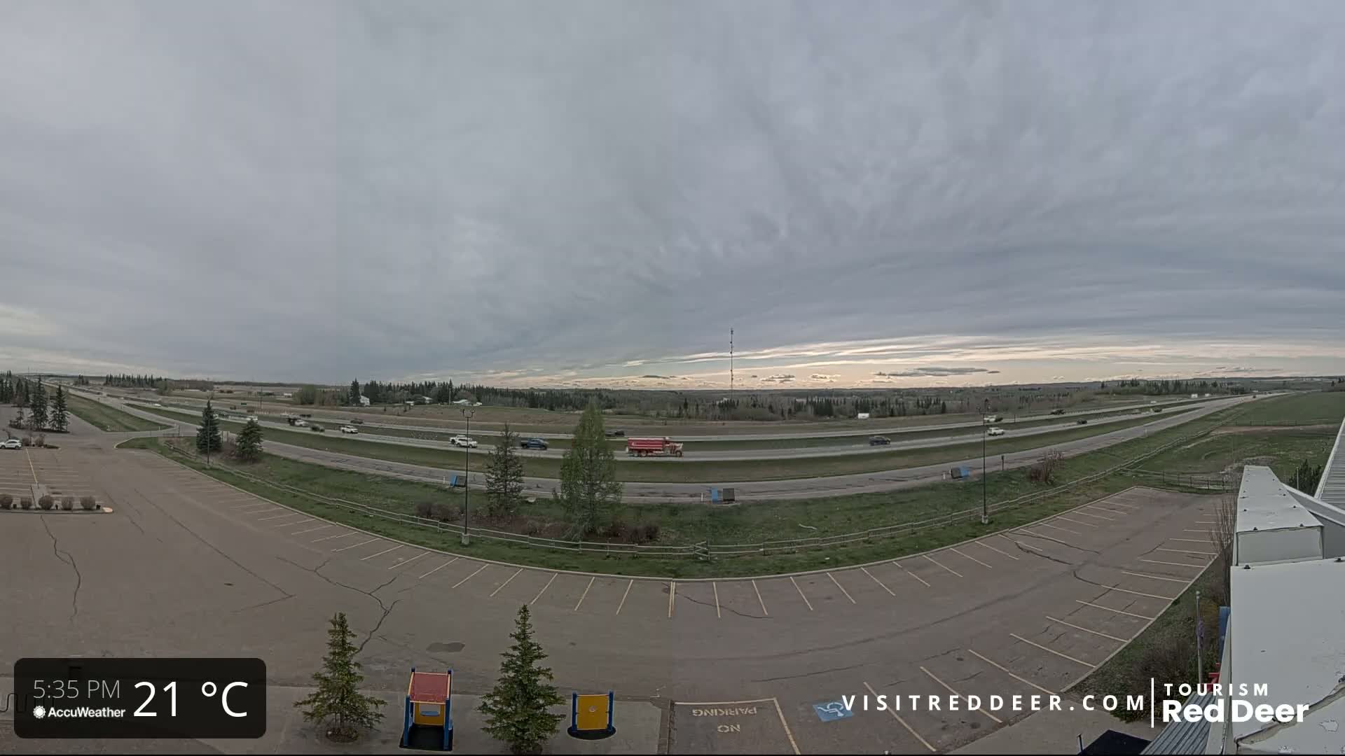 A mostly cloudy sky hangs over a multi-lane highway with sparse traffic and an adjacent empty parking lot.