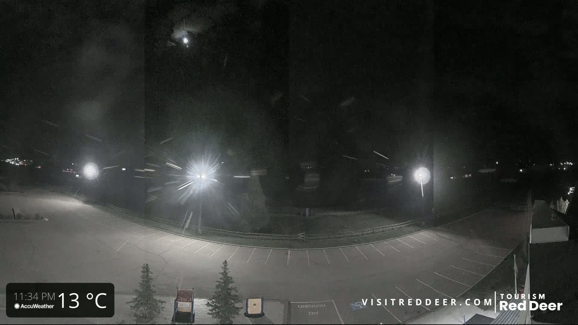 A nighttime view of a snow-covered empty parking lot illuminated by several bright lights under a dark sky with light snow falling.