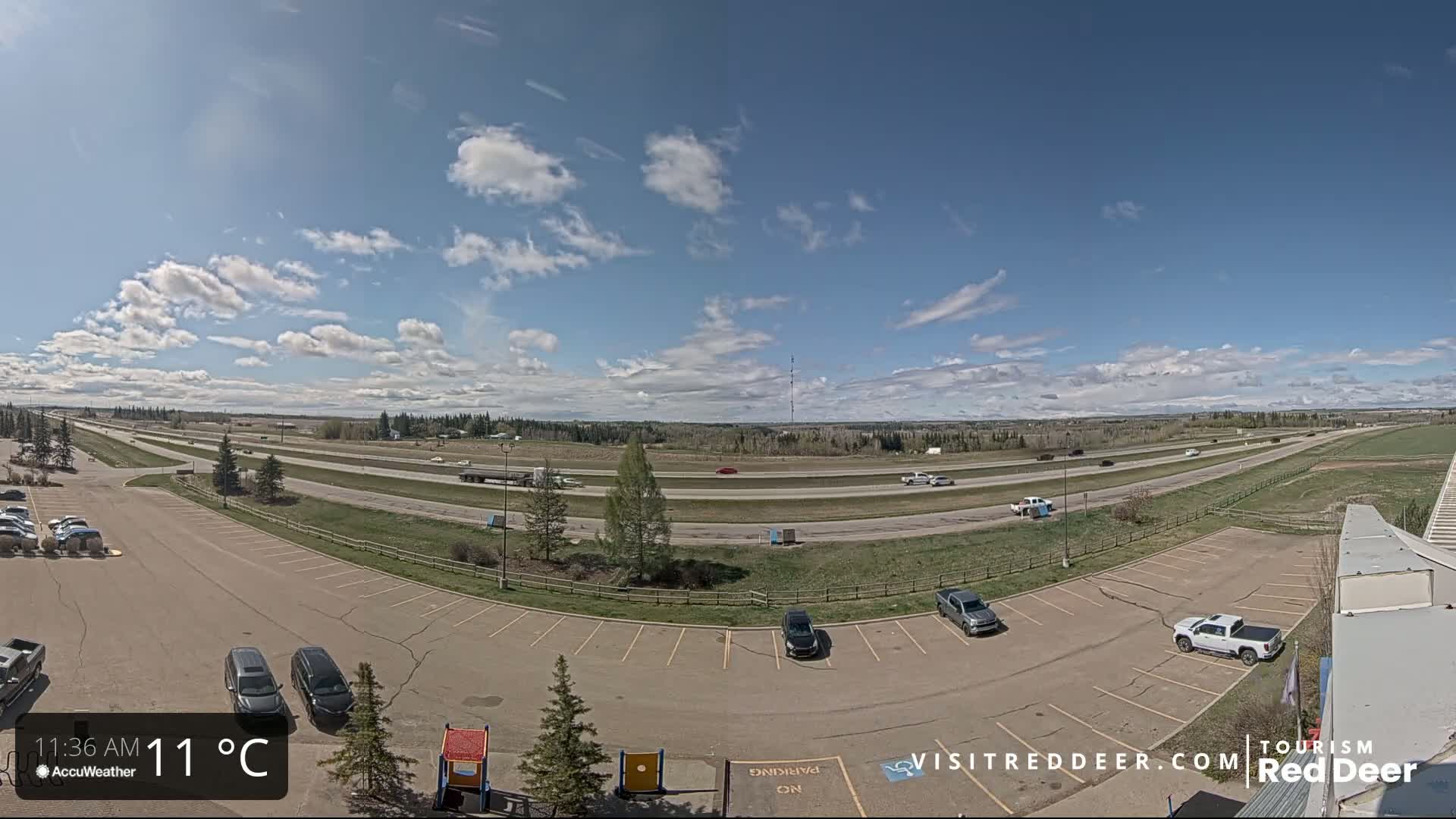 A partly cloudy sky overlooks a highway with several vehicles, adjacent to a parking lot with cars, under a sunny sky with a temperature of 11°C.
