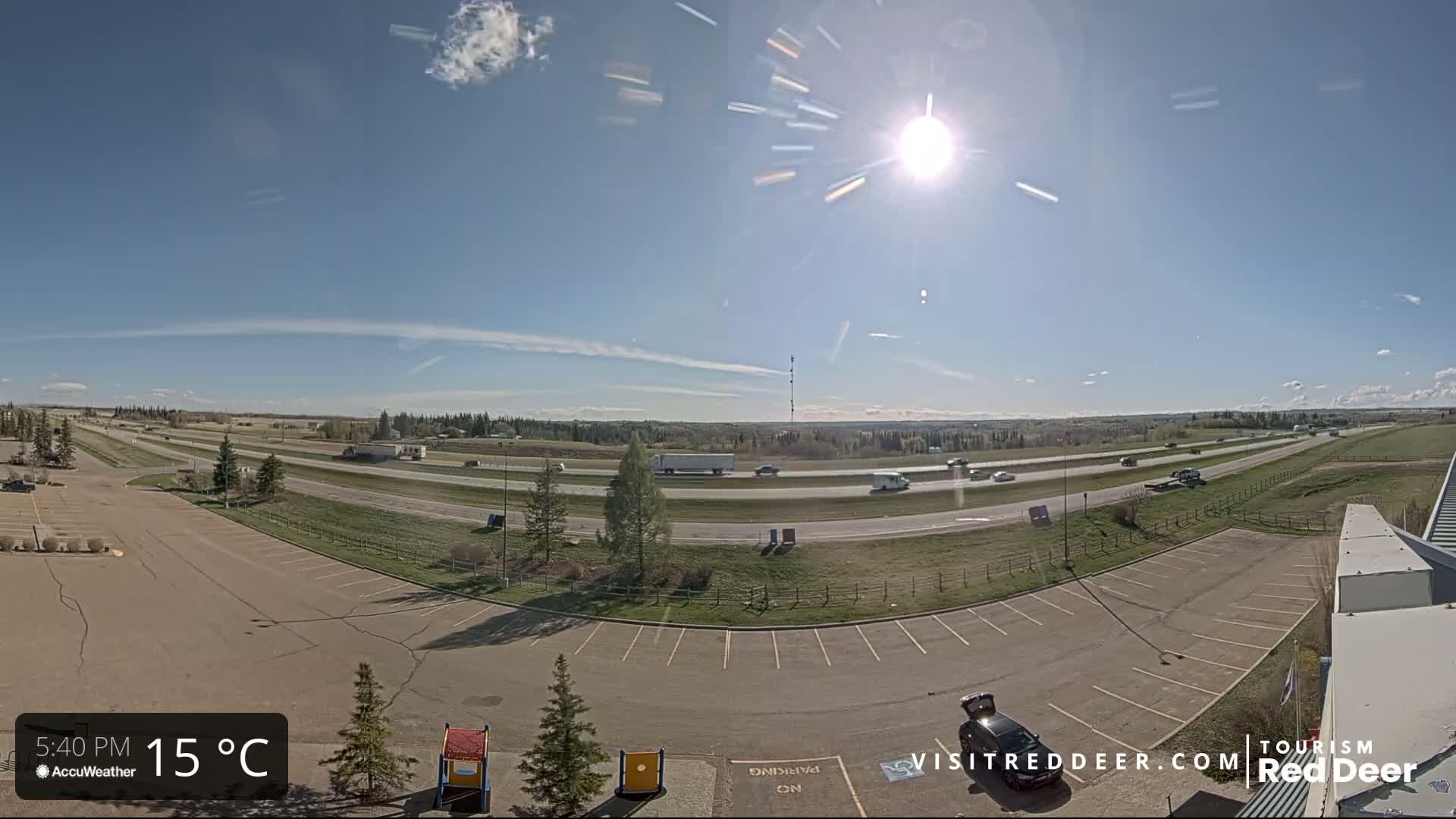 A sunny, mostly clear day reveals a highway with moderate traffic, adjacent to an empty parking lot and a grassy area.