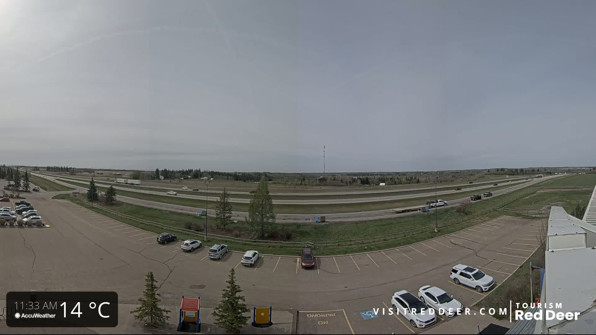 A mostly sunny day reveals a highway with sparse traffic and a parking lot with several cars, surrounded by grassy areas and trees.