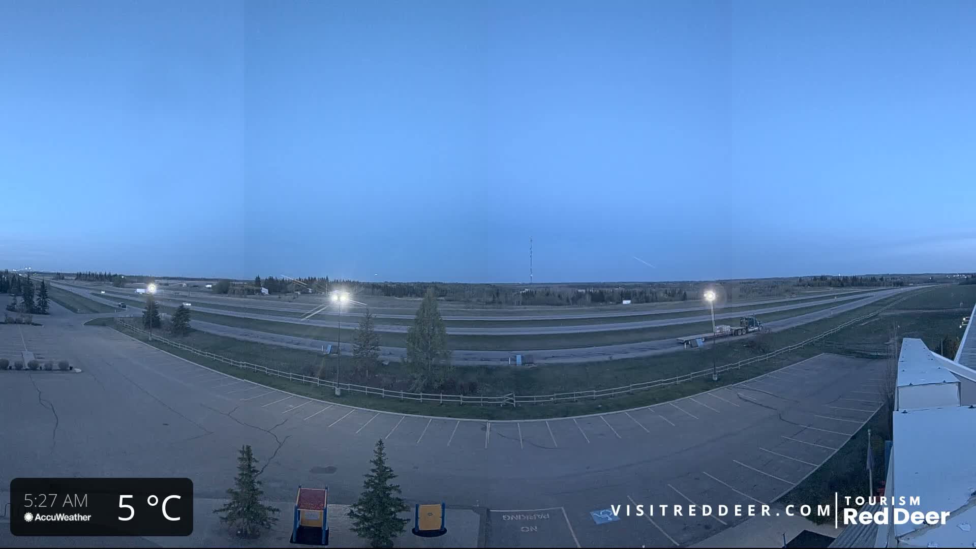 A mostly empty parking lot overlooks a highway and flat, grassy landscape under a twilight sky, with a temperature of 5°C.