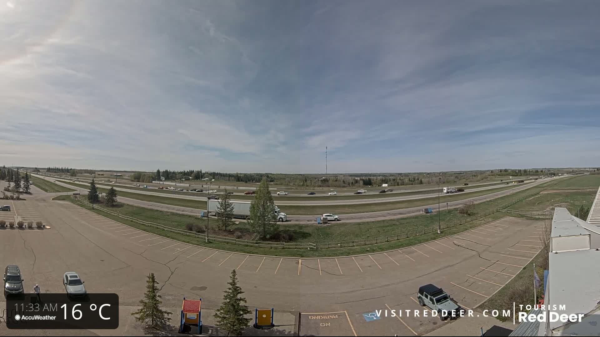 A mostly sunny day shows a highway with moderate traffic, an adjacent parking lot with a few cars, and a grassy area beyond.