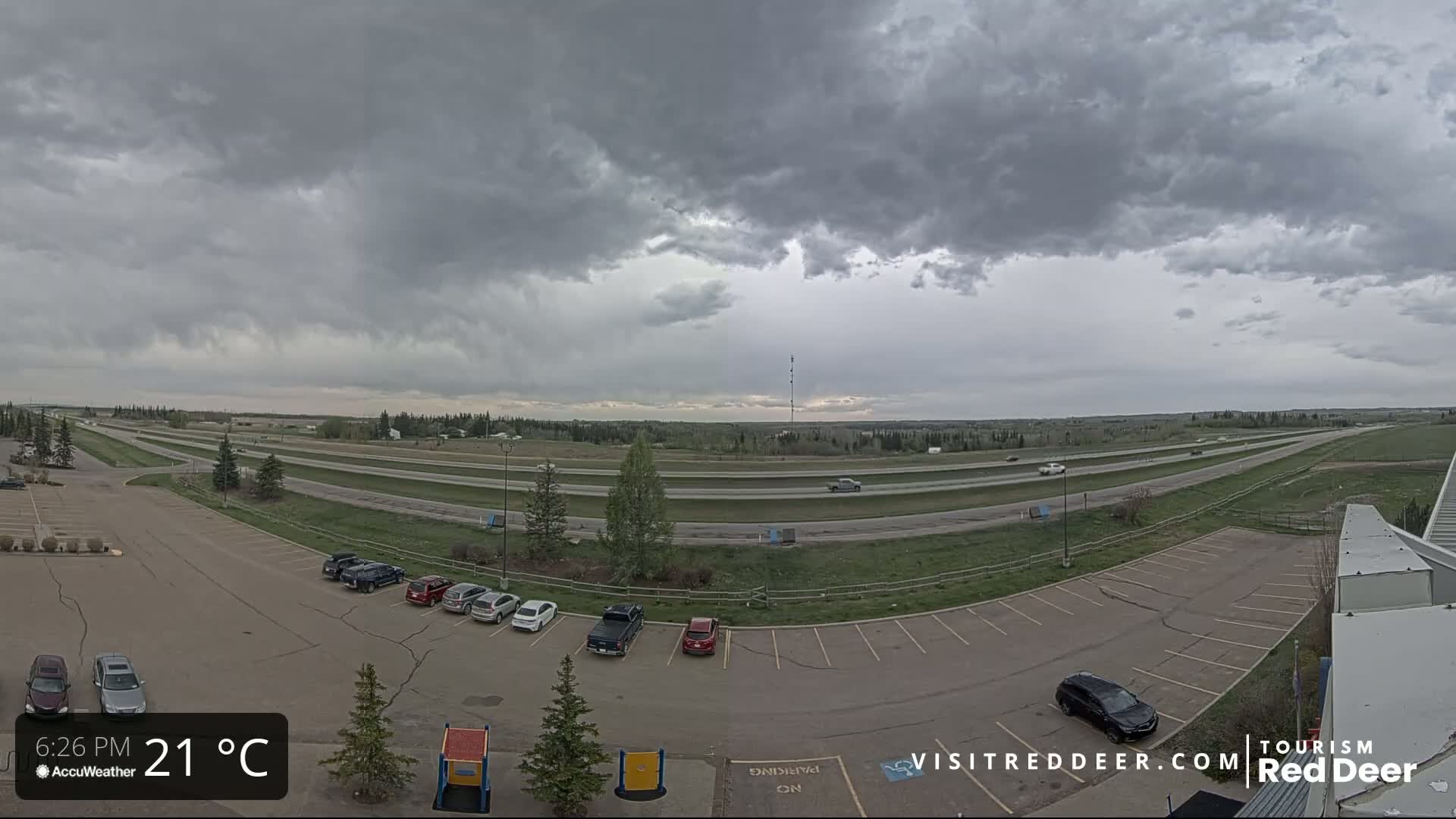A mostly cloudy sky hangs over a highway and a parking lot with several cars, some small trees, and patches of grass.
