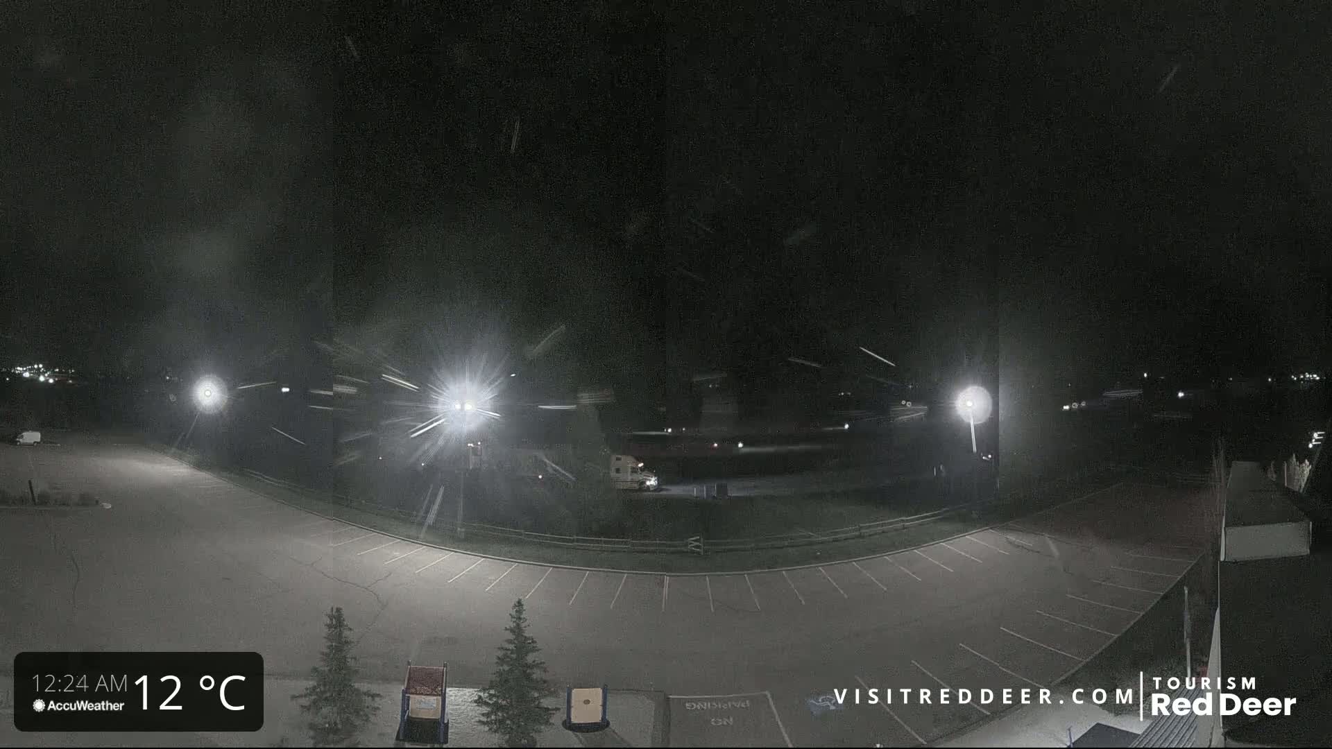 A nighttime, snow-covered parking lot is illuminated by several bright lights, with light snow falling.