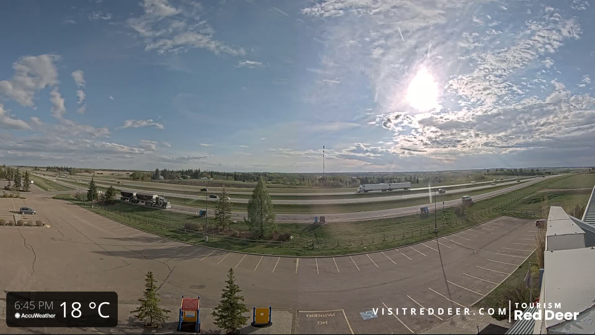 A mostly sunny day with partly cloudy skies shows a highway with several vehicles and an empty parking lot next to a grassy area.