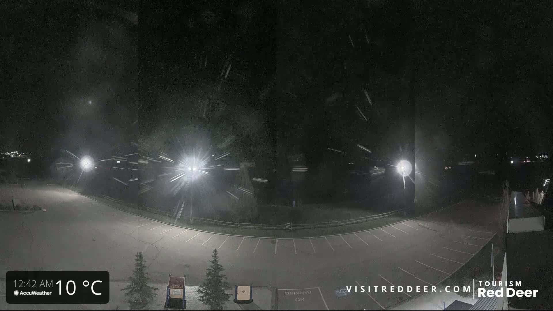 Snow is falling at night over an empty parking lot illuminated by several bright lights.