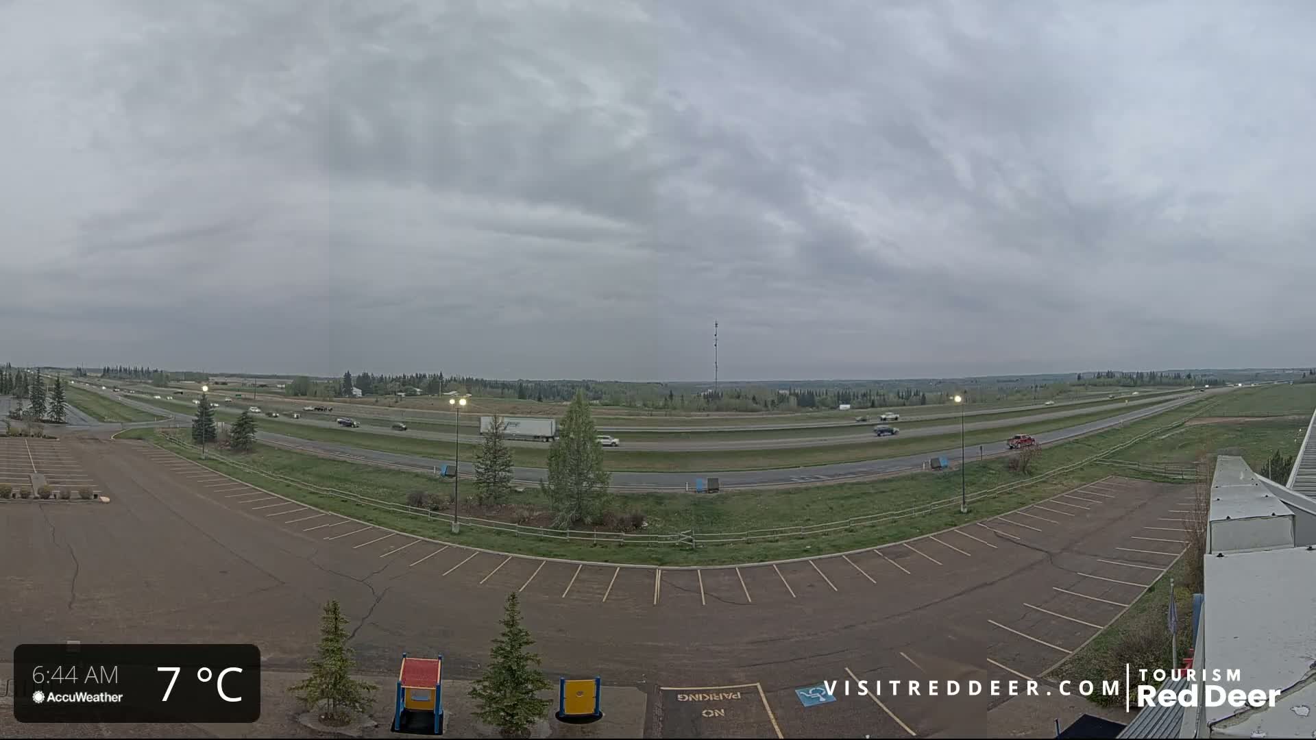 An overcast sky hangs over a highway with sparse traffic and an empty parking lot next to a grassy area.