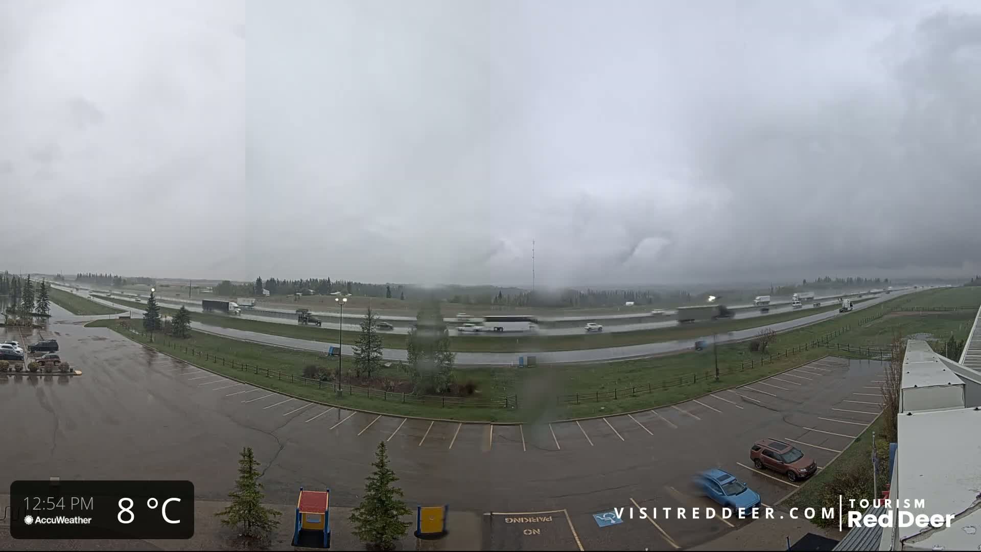 A wet parking lot overlooks a highway with light traffic under a gray, overcast sky.