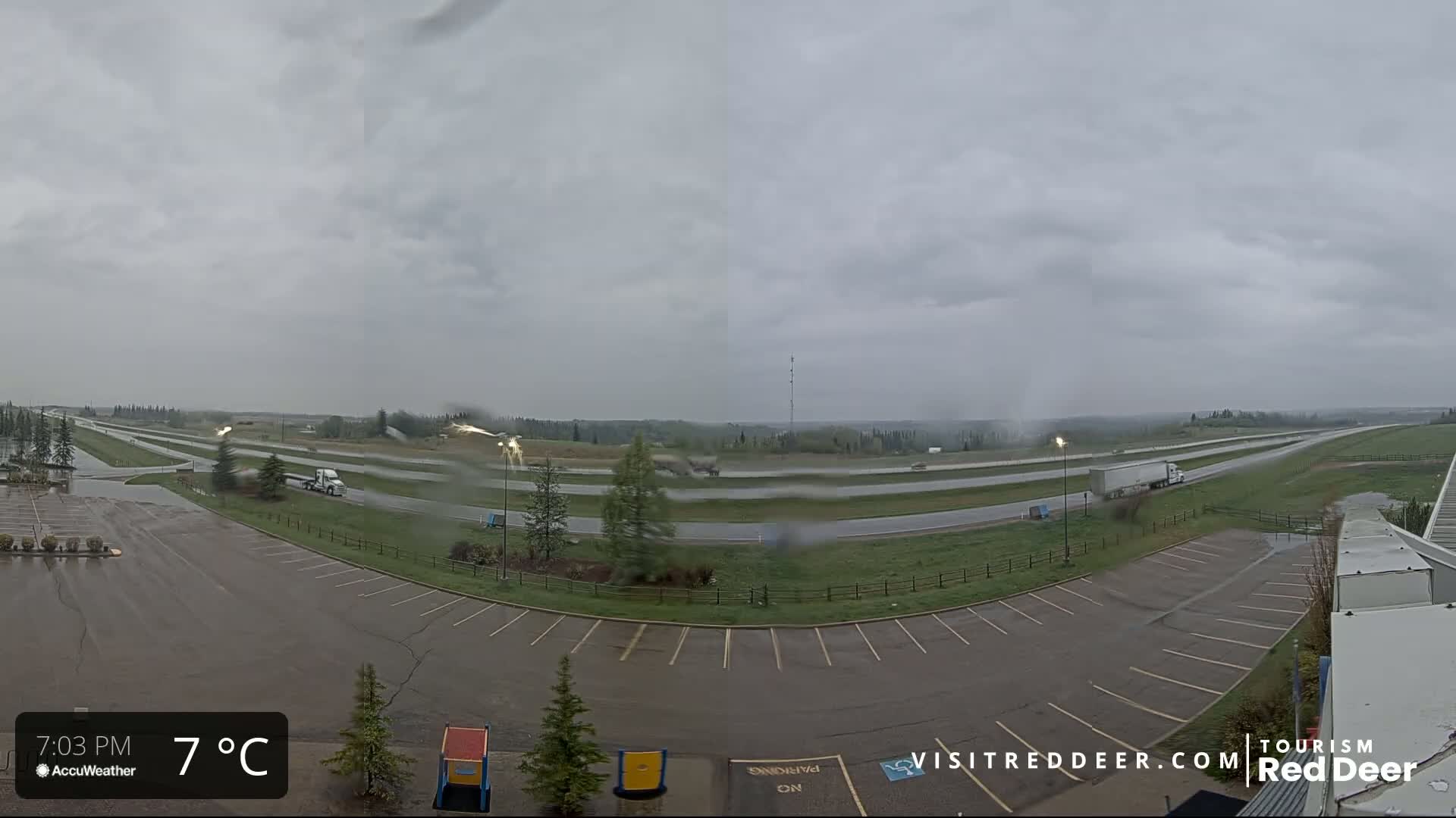 A wet parking lot and highway are visible under a gray, overcast sky, with a few vehicles traveling on the highway.