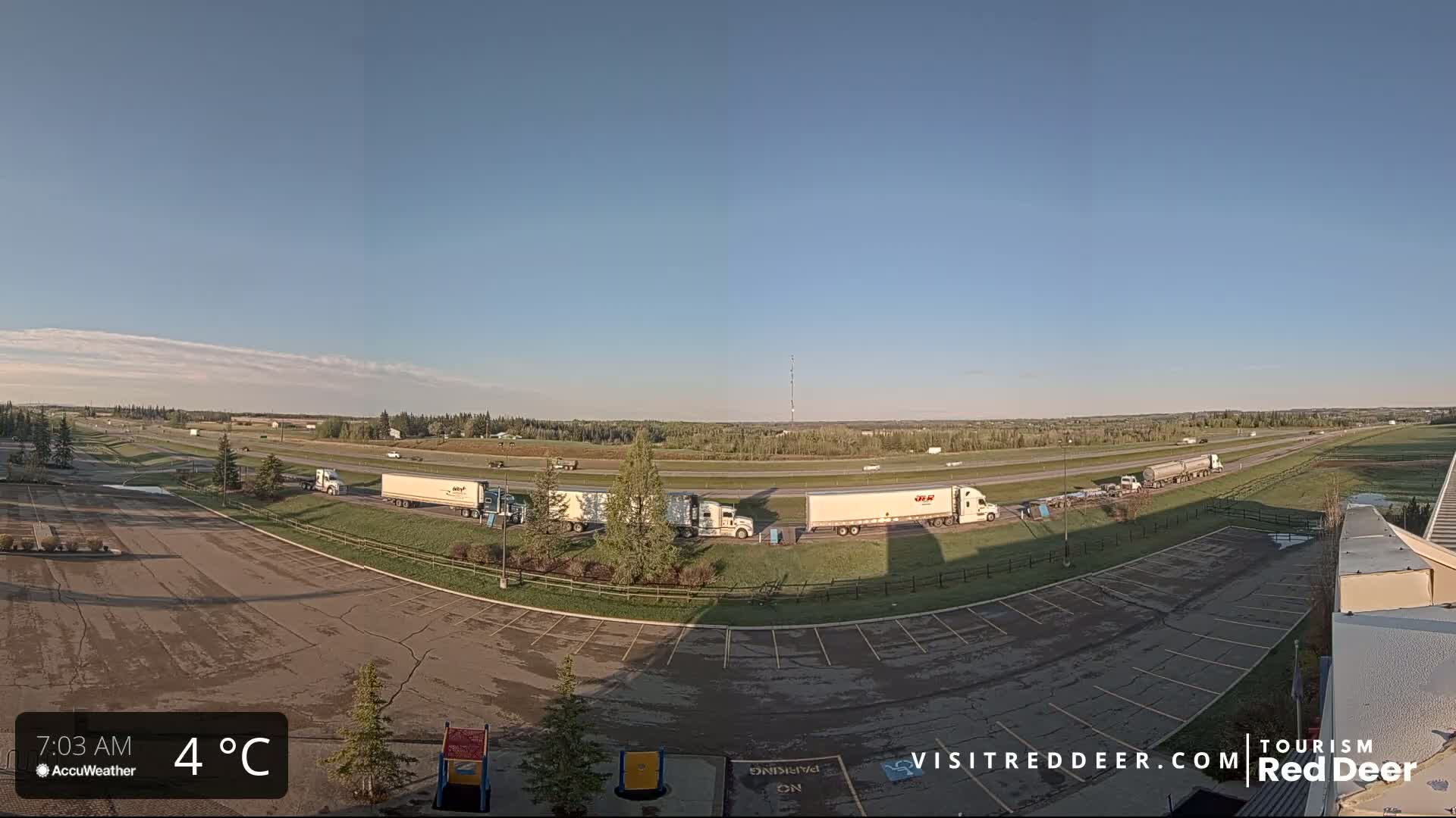 A mostly empty parking lot next to a highway with several semi-trucks stopped along the side of the road on a partly sunny, cool day.
