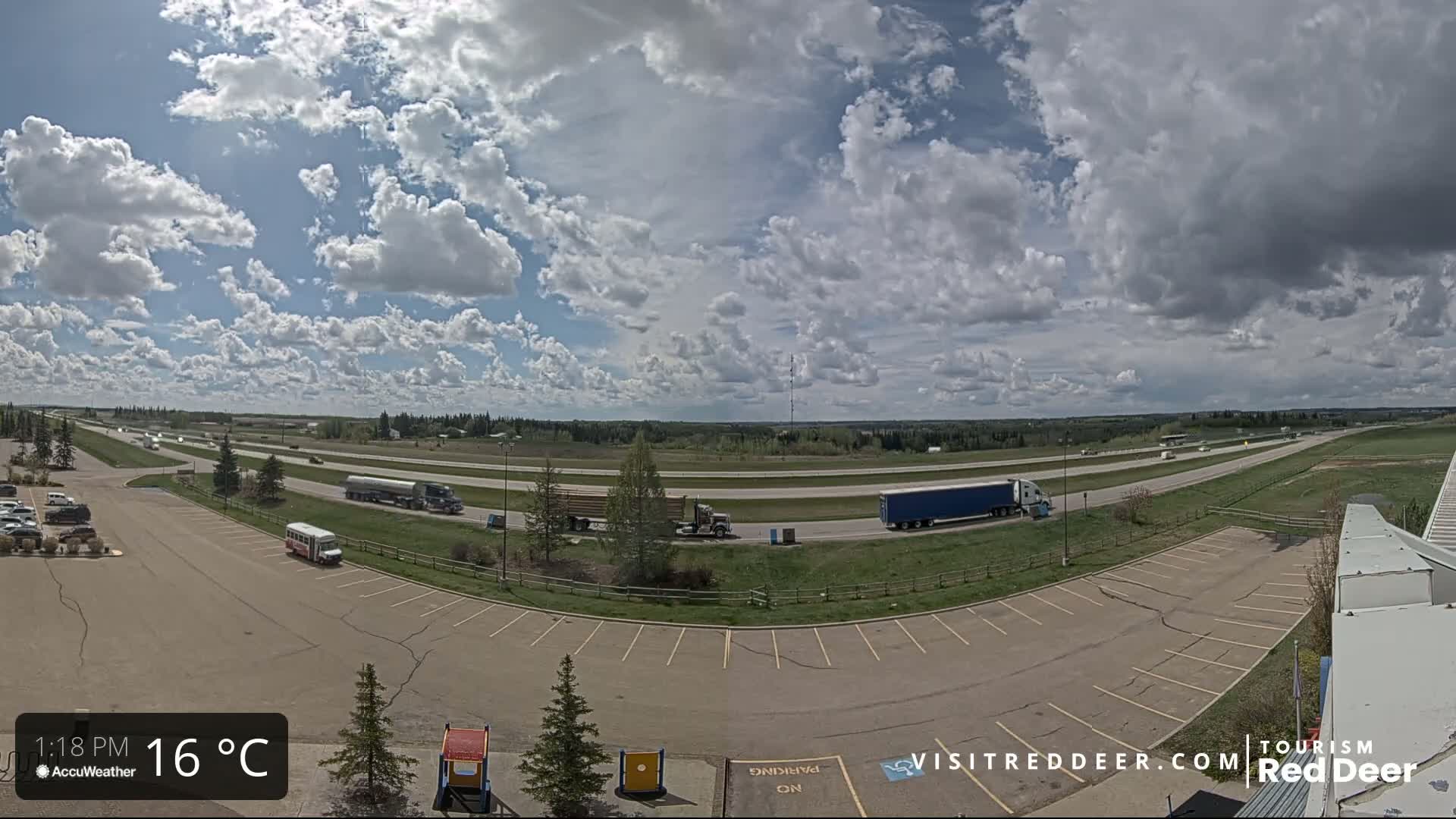 A mostly sunny day with partly cloudy skies shows a highway with several vehicles, including semi-trucks, and a large parking lot with a few cars and a bus.