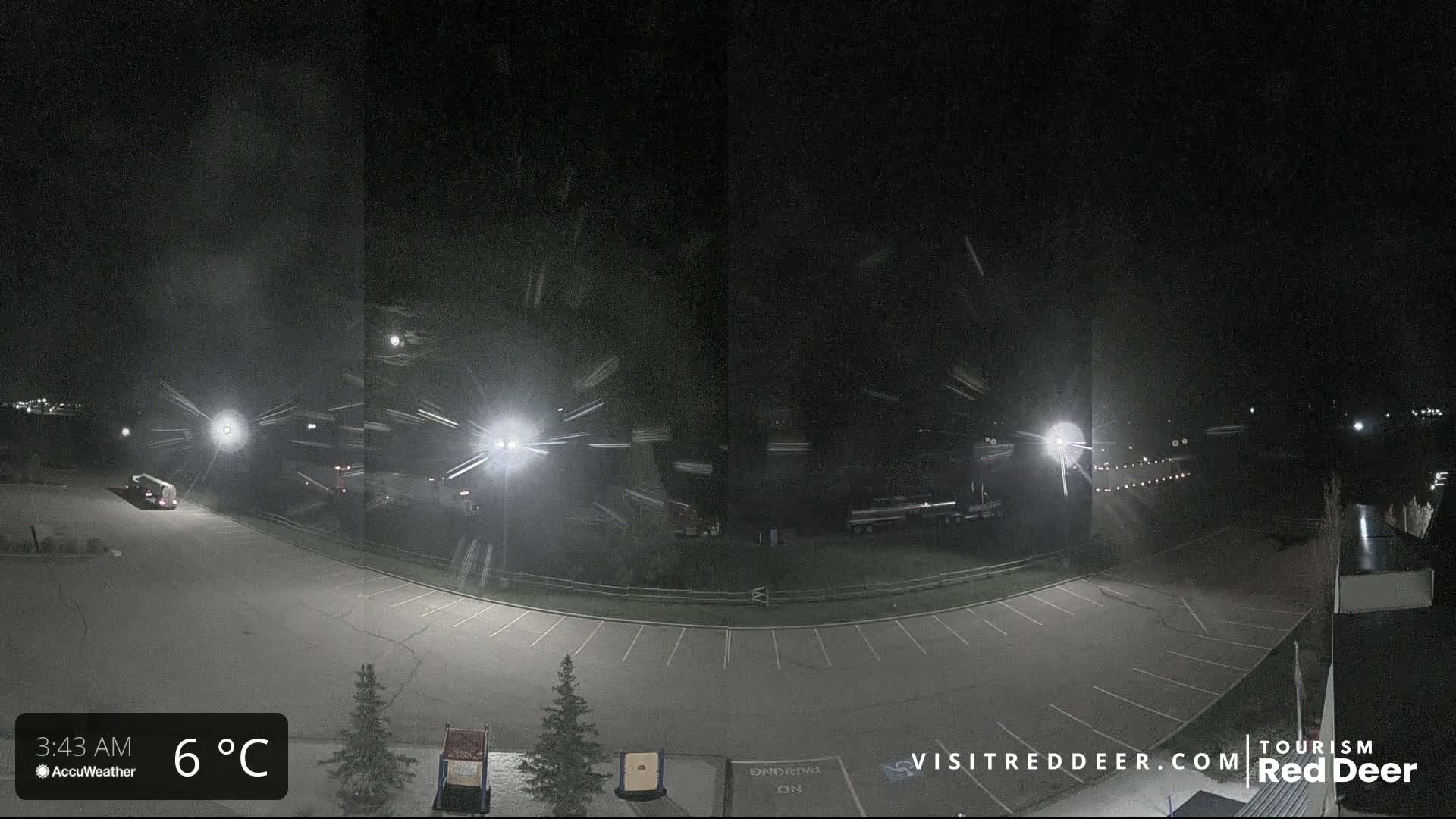 A nighttime scene of a mostly empty parking lot with several vehicles and light poles, under snowy conditions.