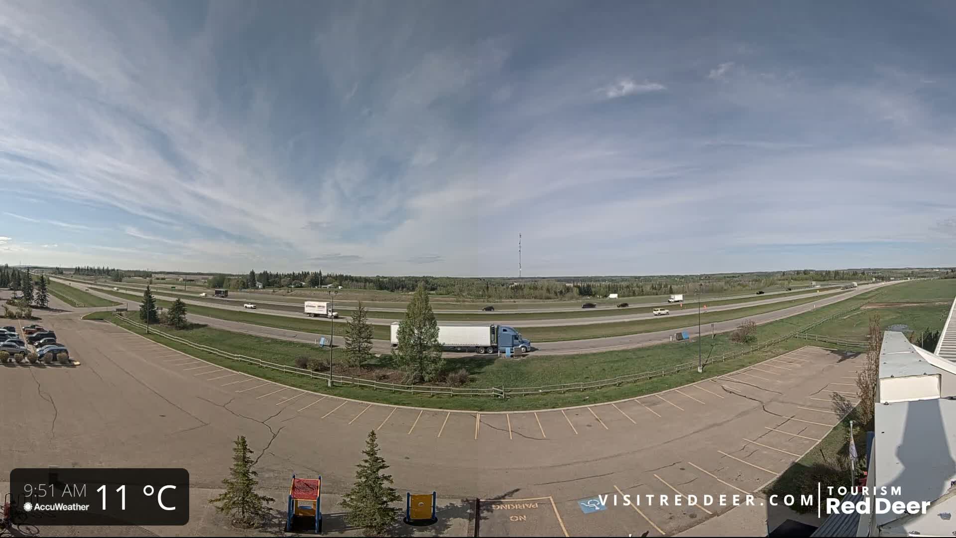 A partly cloudy sky overlooks a highway with several vehicles, including semi-trucks, and an adjacent empty parking lot.