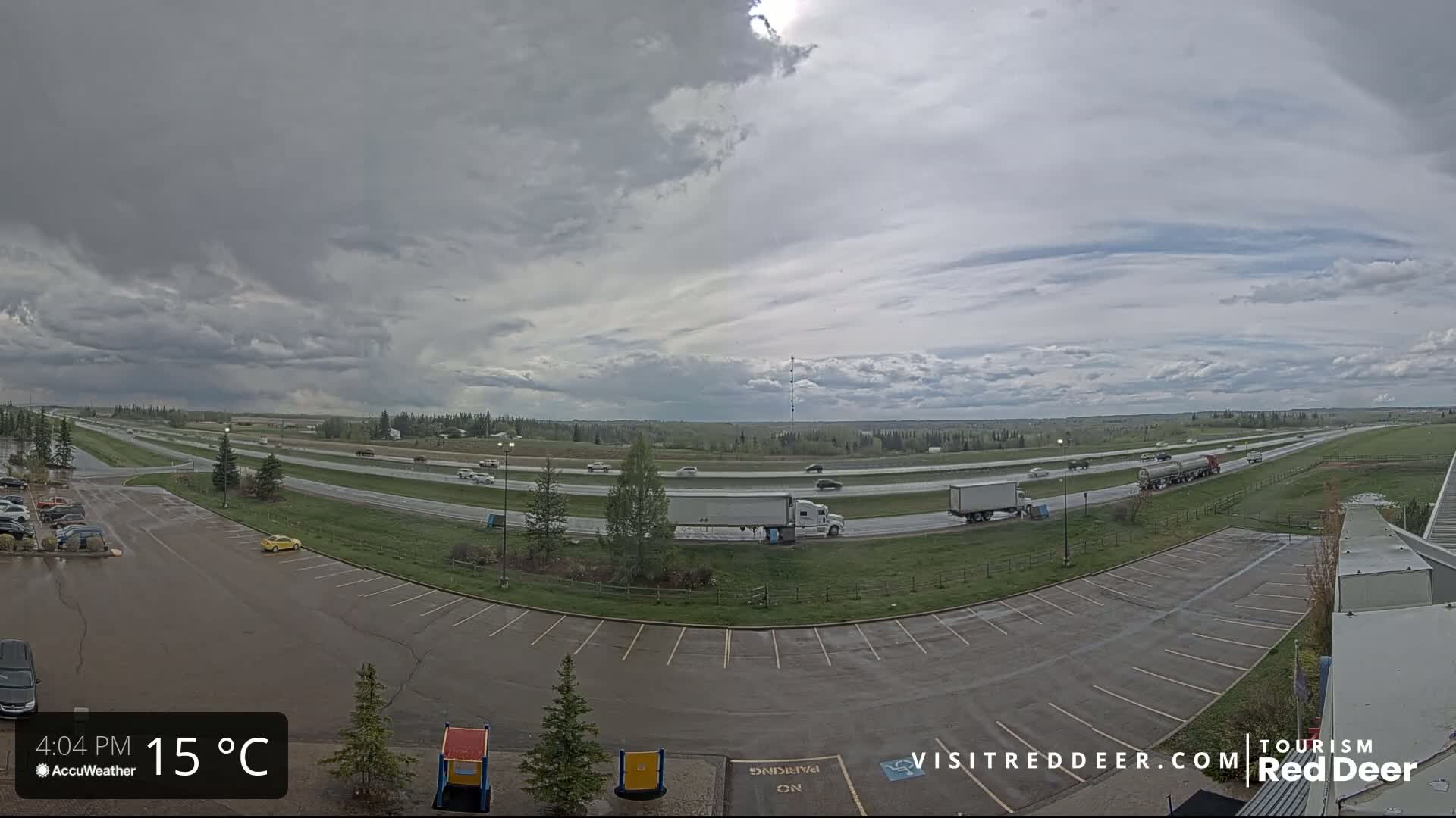 A mostly cloudy sky hangs over a highway with moderate traffic and a large, empty parking lot, viewed from an elevated perspective.