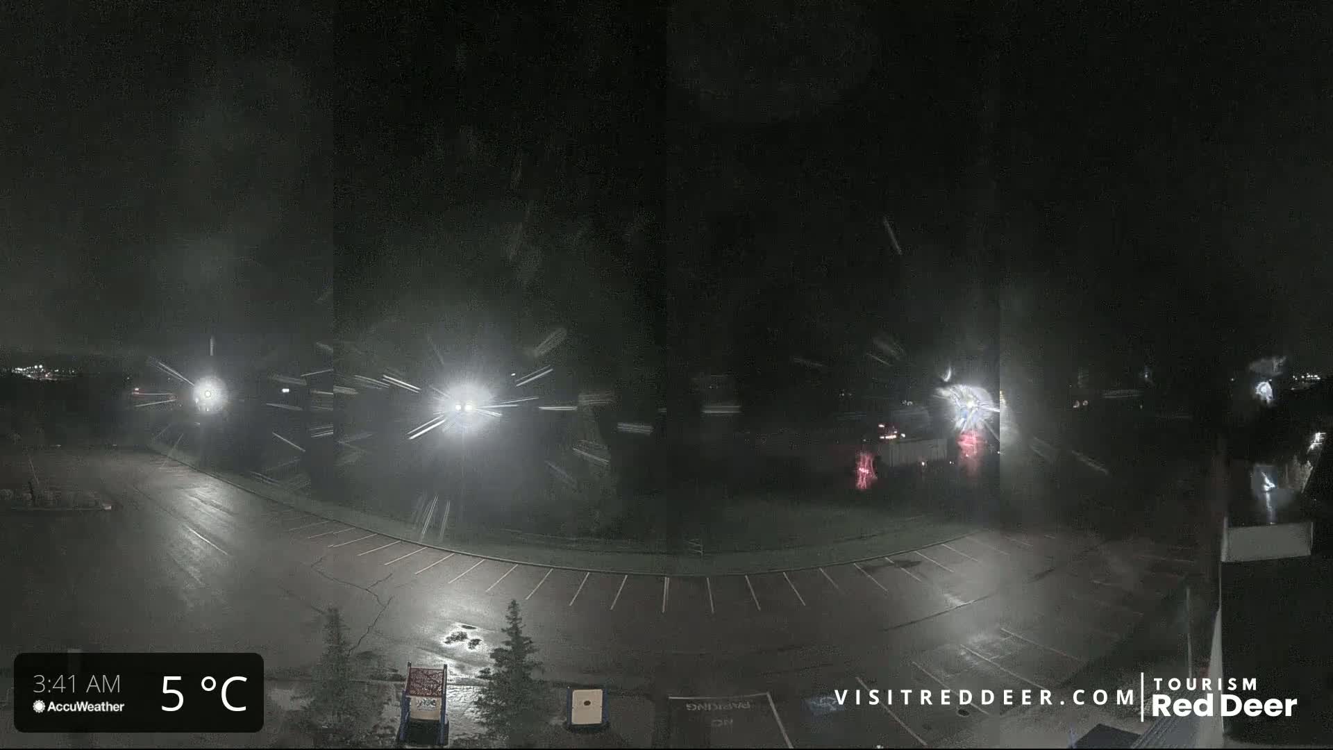 A nighttime scene of a snow-covered parking lot with streetlights illuminating falling snow and a few distant vehicles.