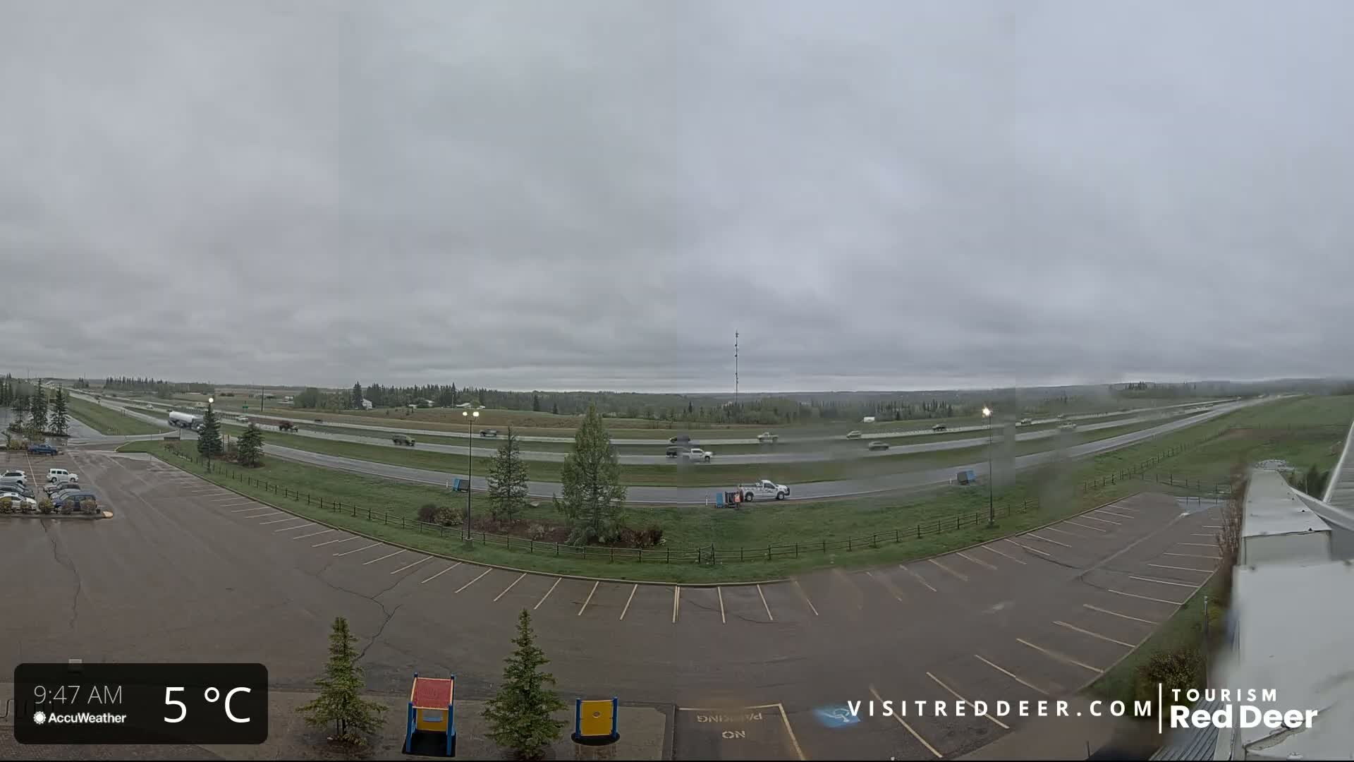 A mostly empty parking lot overlooks a highway with light traffic under an overcast sky, lightly sprinkling rain.