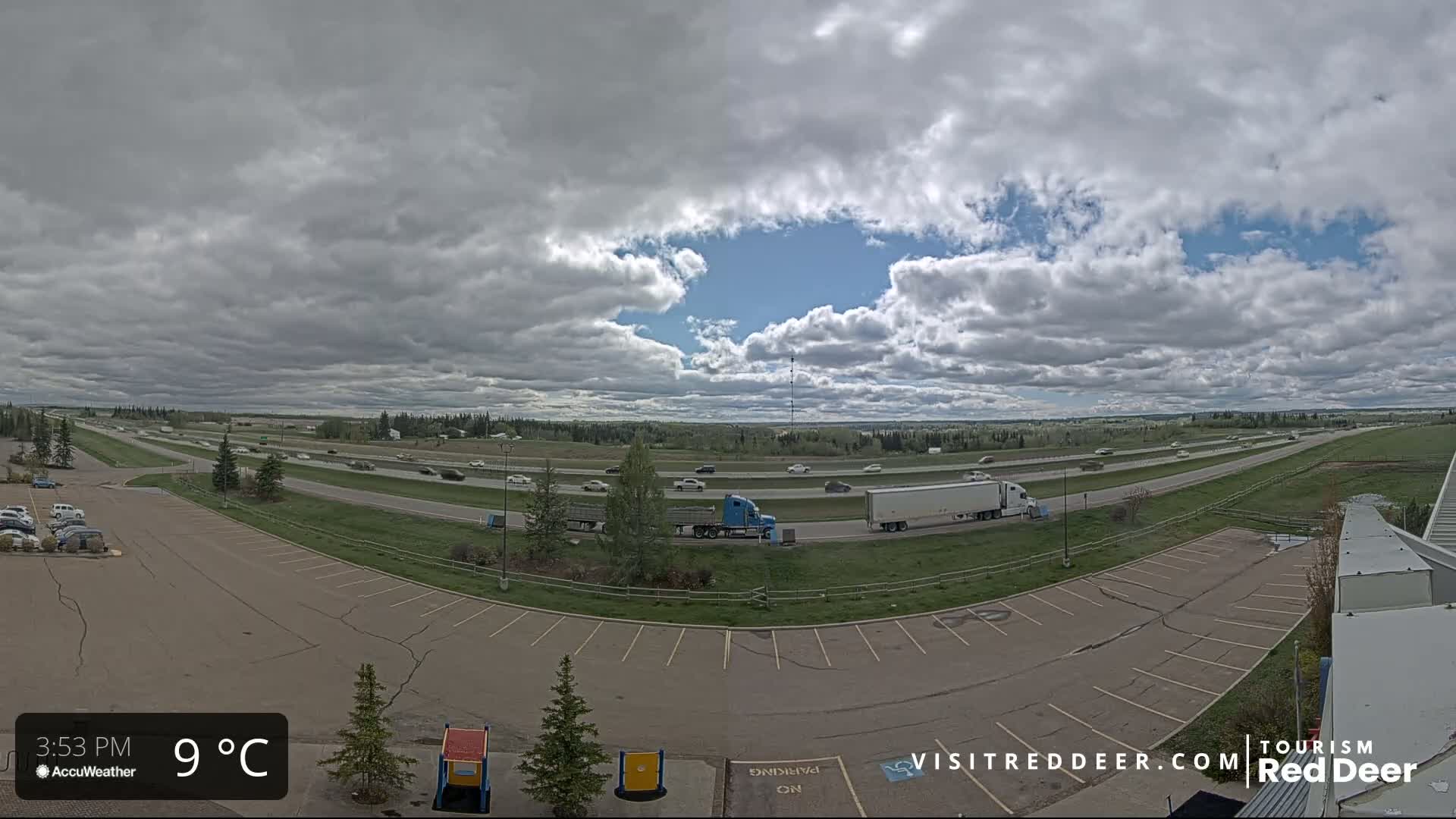 A mostly empty parking lot overlooks a highway with light traffic under an overcast sky, lightly sprinkling rain.