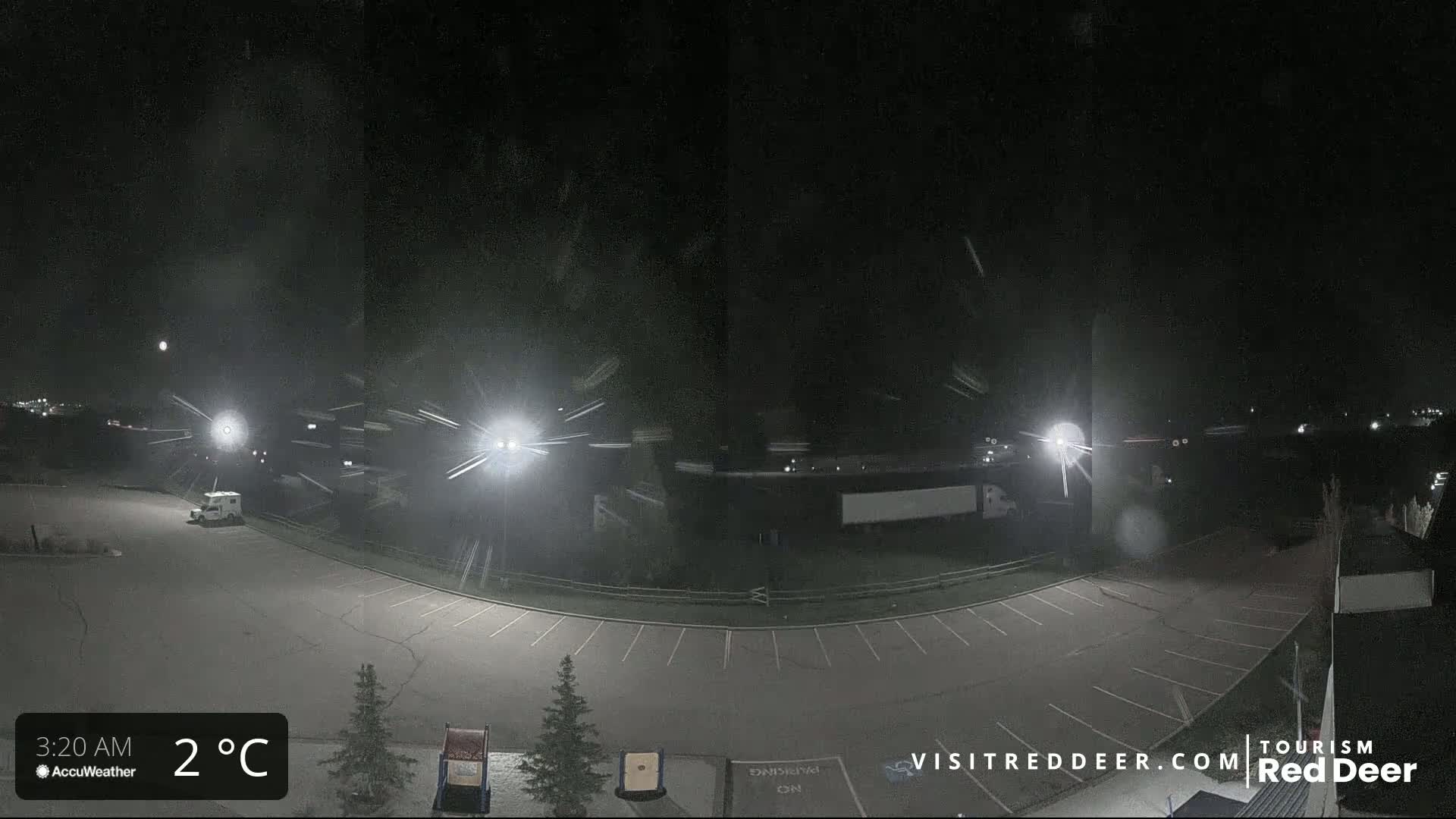 A nighttime, snow-covered parking lot with a few vehicles is illuminated by bright lights under a dark sky.