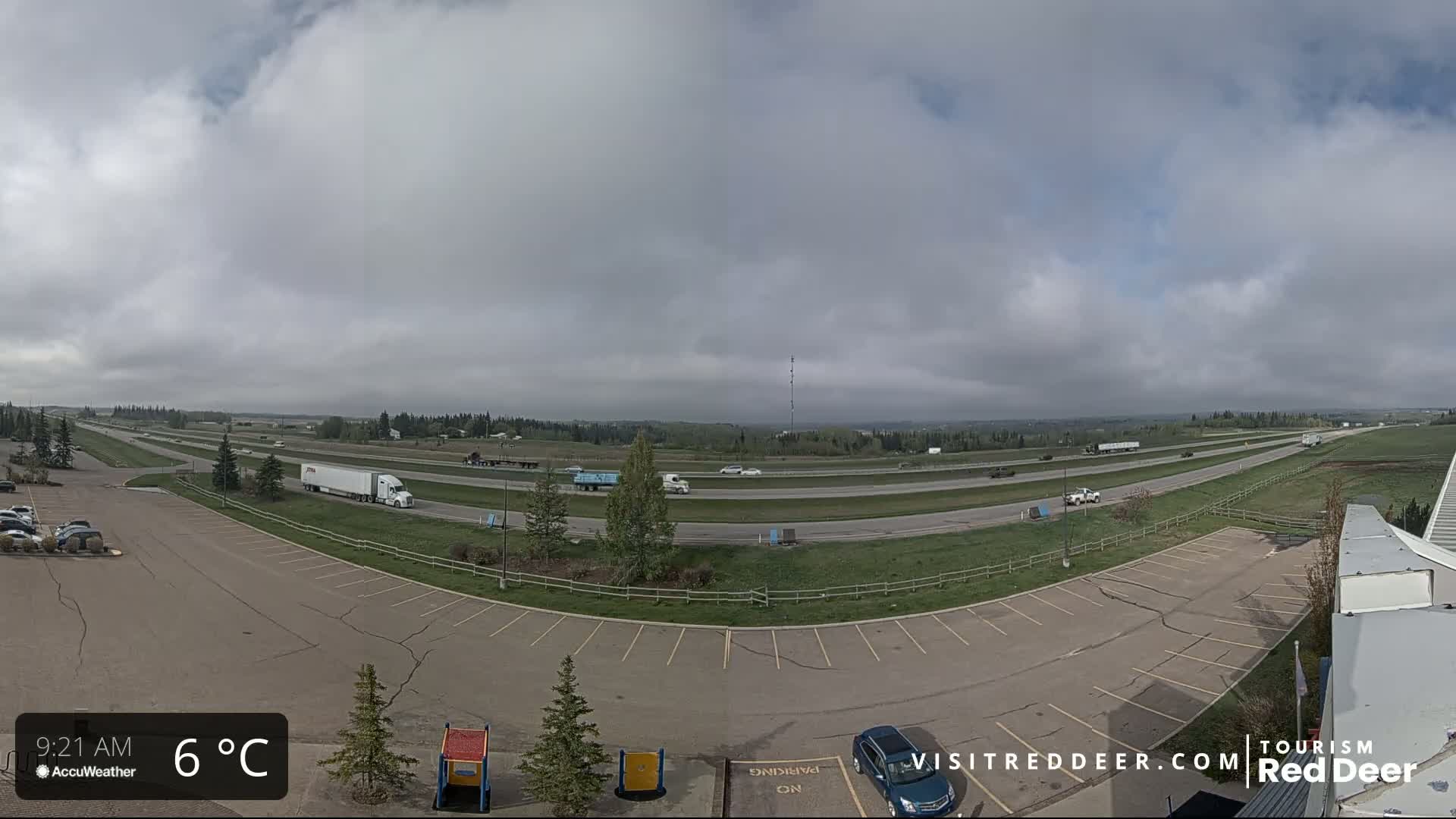A mostly cloudy sky hangs over a highway with several vehicles, including semi-trucks, and a mostly empty parking lot.