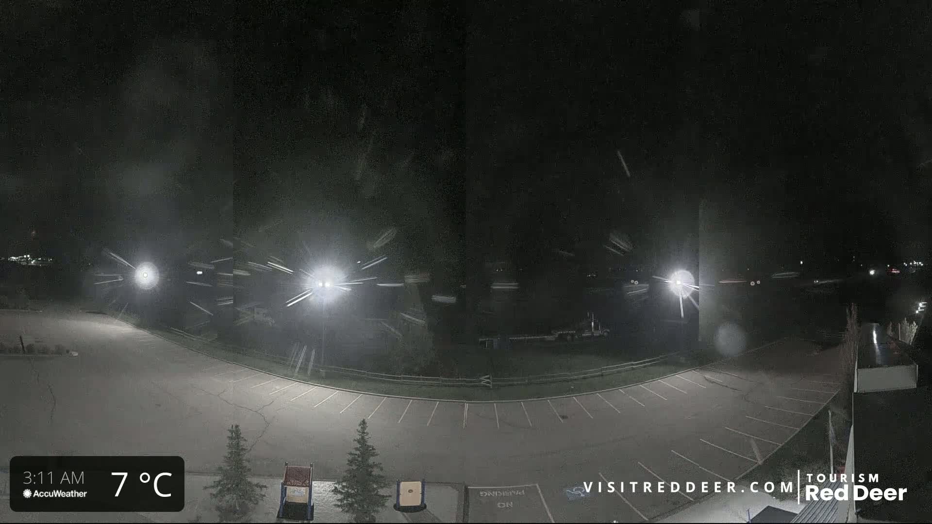 A nighttime, snow-covered parking lot is illuminated by several bright lights under a dark, snowy sky.
