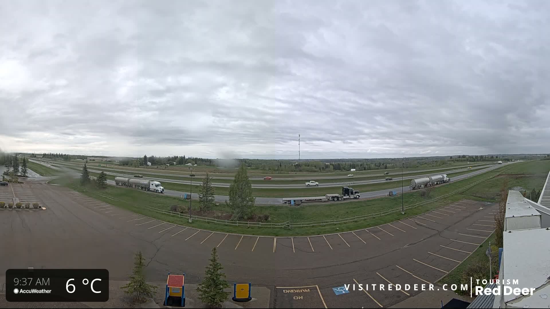 A mostly empty parking lot overlooks a highway with several vehicles and semi-trucks under an overcast sky.