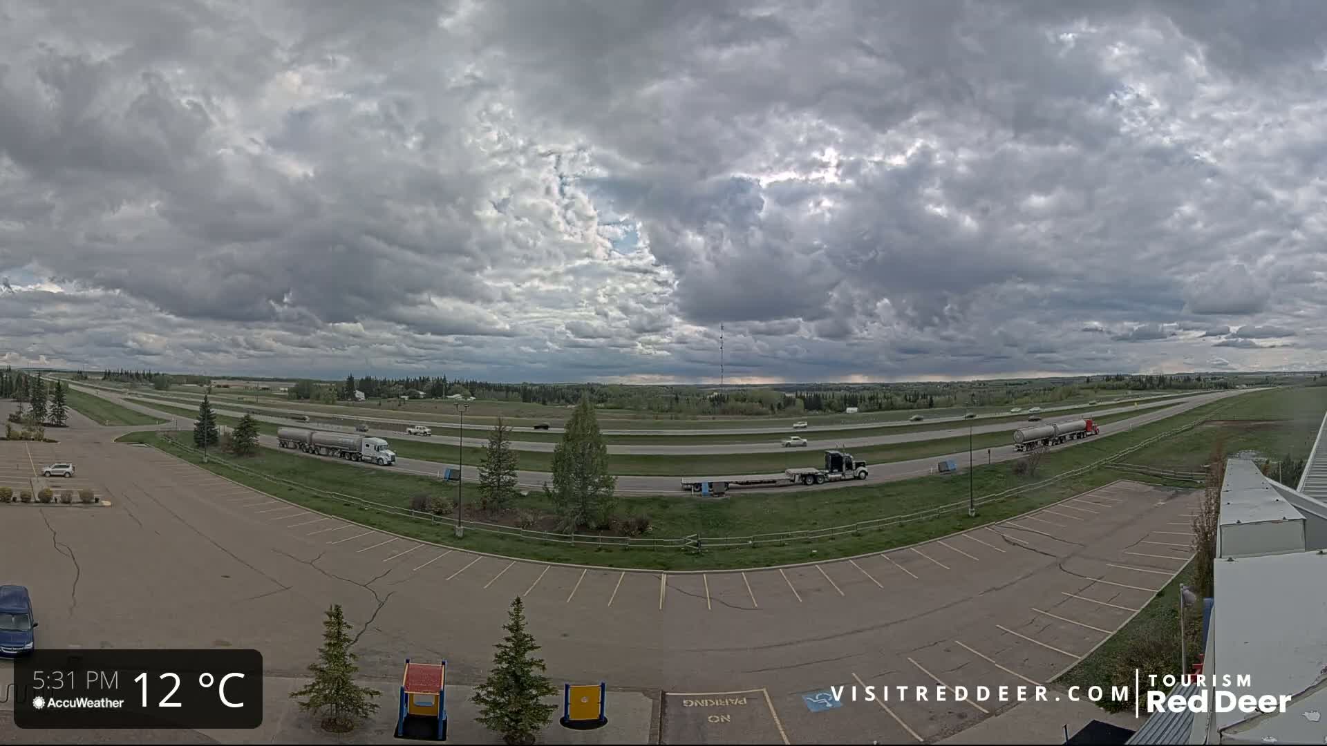 A mostly cloudy sky hangs over a highway with several semi-trucks and cars, as seen from a parking lot with some vehicles parked.