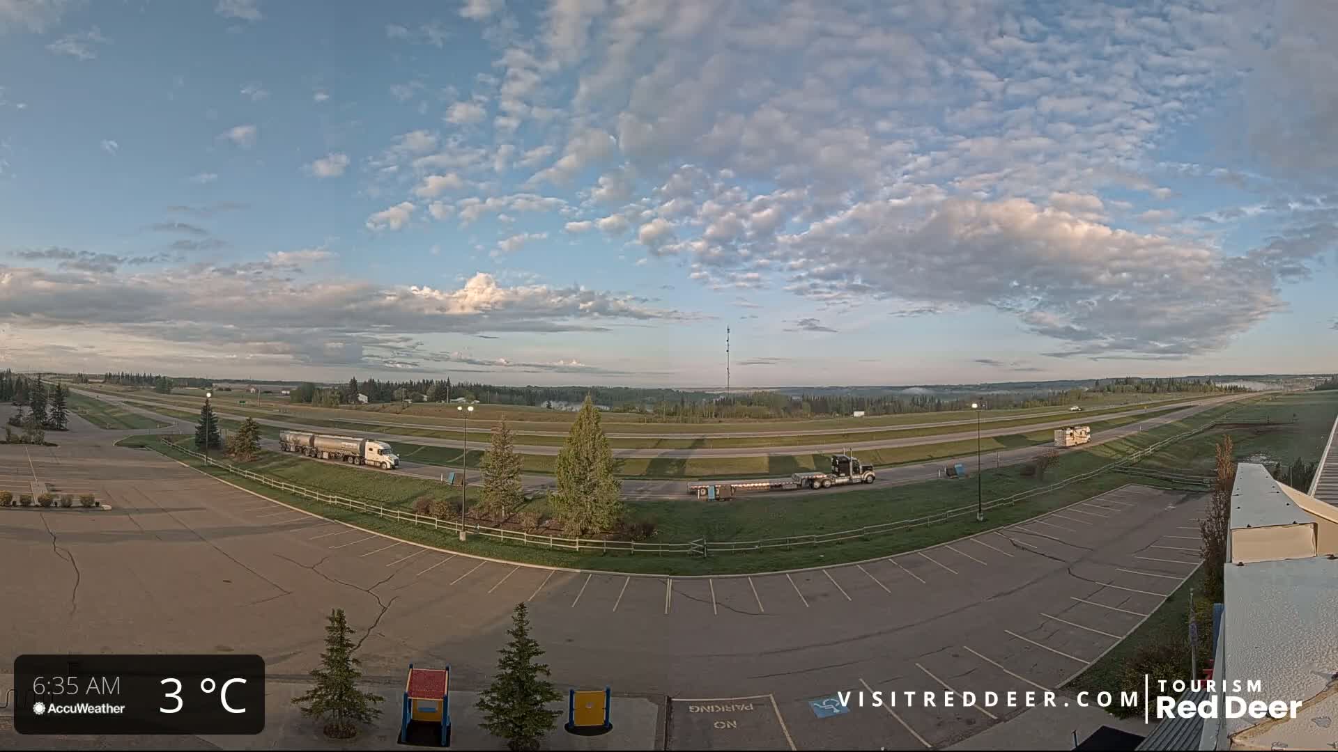 An empty parking lot overlooks a highway with several trucks, under a partly cloudy sky, with a temperature of 3°C.