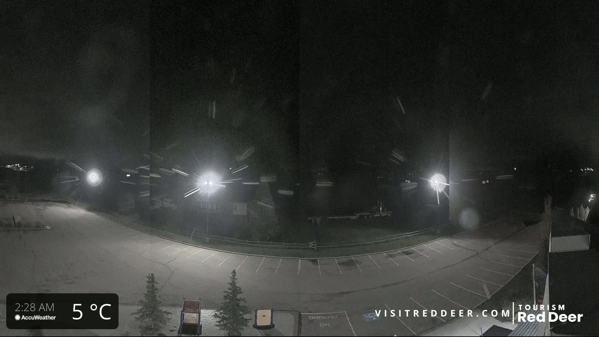 A nighttime view of a mostly empty parking lot with light snow falling under bright lights.