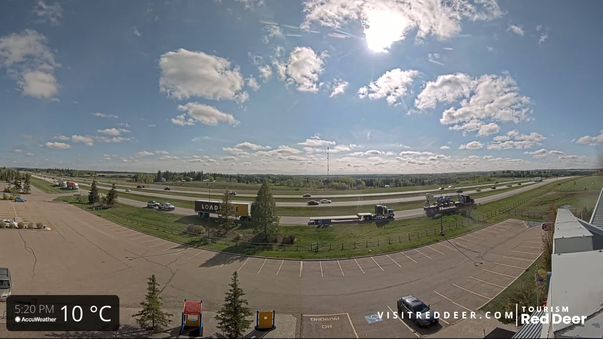 A partly cloudy sunny day shows a highway with several vehicles and an empty parking lot.
