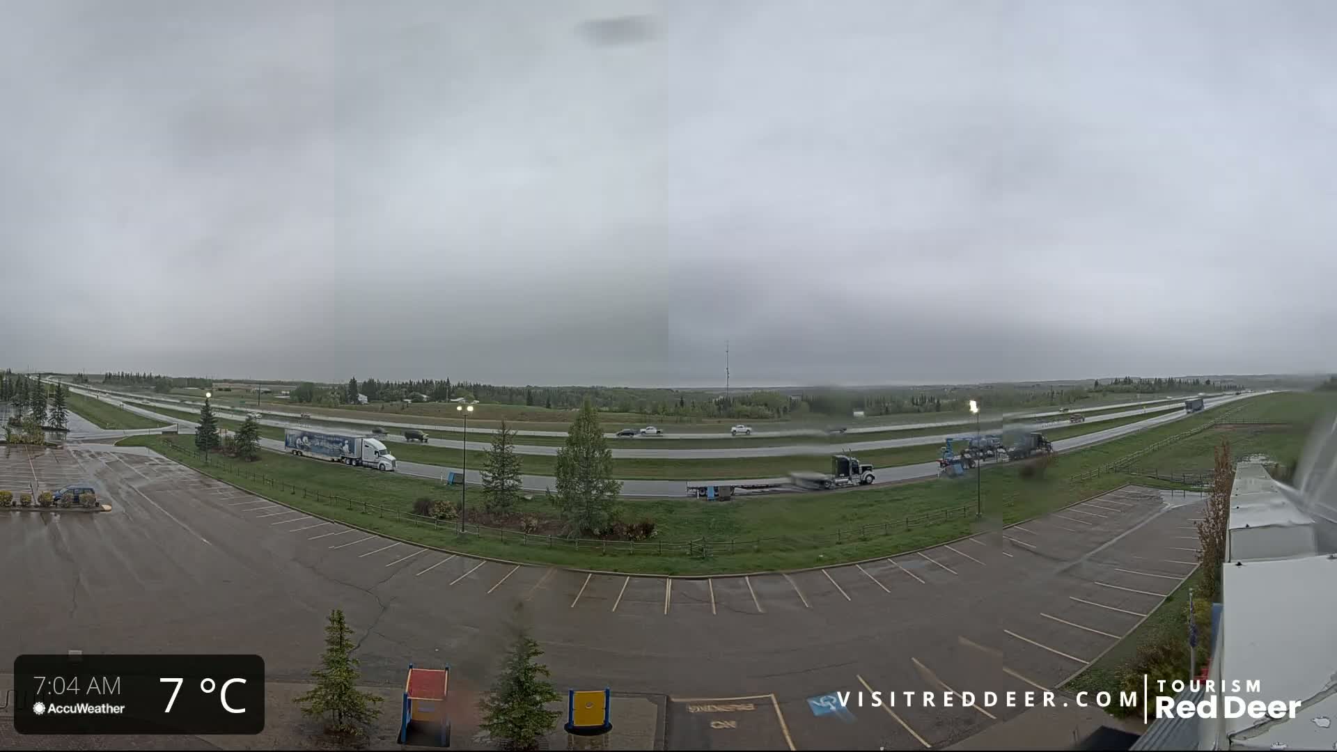A wet, overcast scene shows a highway with several vehicles, including semi-trucks, and an adjacent empty parking lot.
