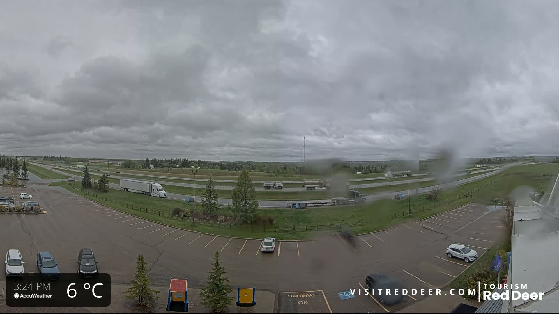A mostly cloudy, rainy day shows a highway with several vehicles and a parking lot with several cars, all surrounded by green grass and trees.